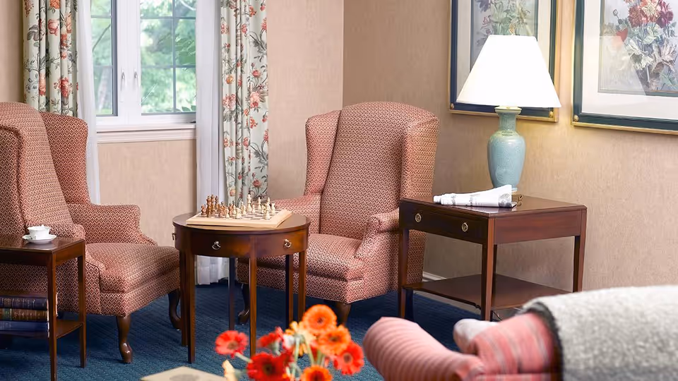 A cozy sitting area with two patterned armchairs facing each other across a small round wooden table with a chess set on it. There is a side table with a lamp and folded napkins, floral curtains framing a window, and framed floral artwork on the wall. A bouquet of red and orange flowers is partially visible in the foreground.