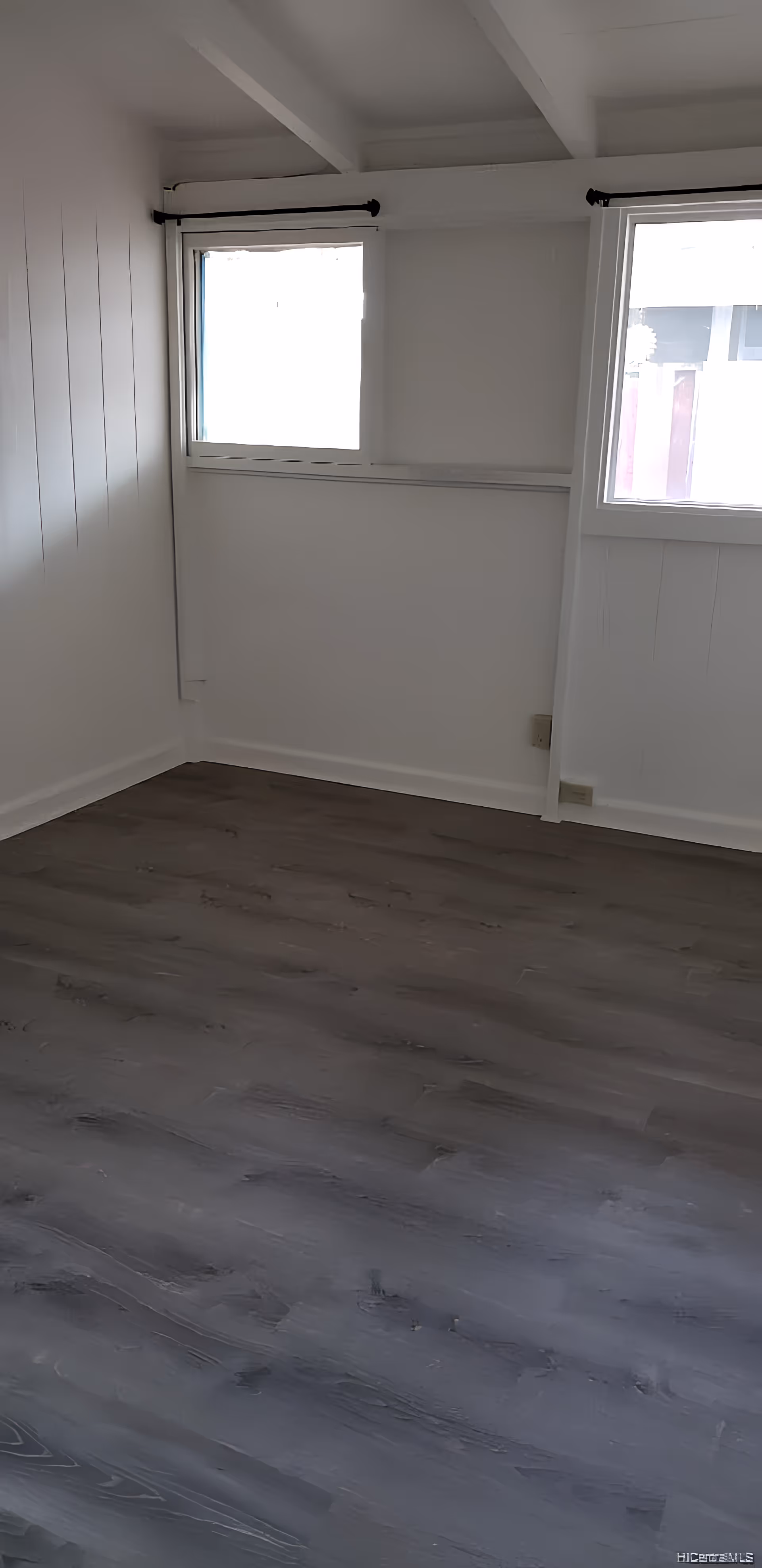 Empty room with white walls, two windows with black curtain rods, and dark wood flooring.