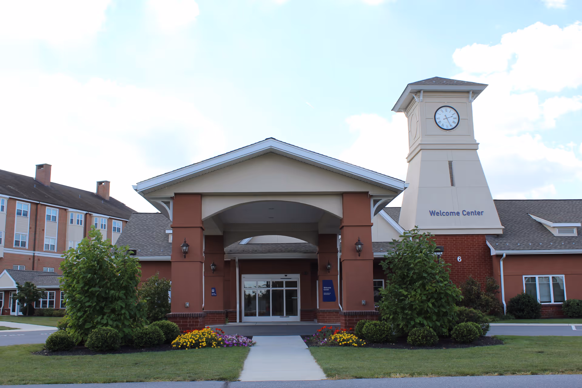 Front entrance of a brick senior living building with a covered portico, landscaping, and a clock tower labeled 'Welcome Center'.