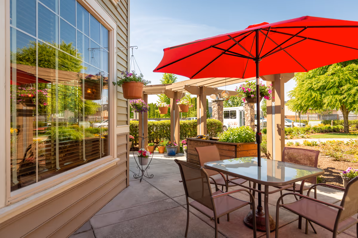 Outdoor patio area with a glass table and four chairs under a large red umbrella. There are hanging flower pots and a wooden pergola with more plants. The patio is adjacent to a building with large windows reflecting greenery and flowers. Trees and shrubs are visible in the background under a clear blue sky.