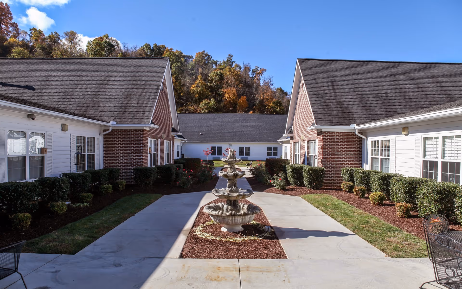 Outdoor courtyard area of a senior living facility with a three-tiered stone fountain in the center, surrounded by neatly trimmed bushes and two single-story buildings with brick and white siding. The sky is clear and blue, and there are trees with autumn foliage in the background.