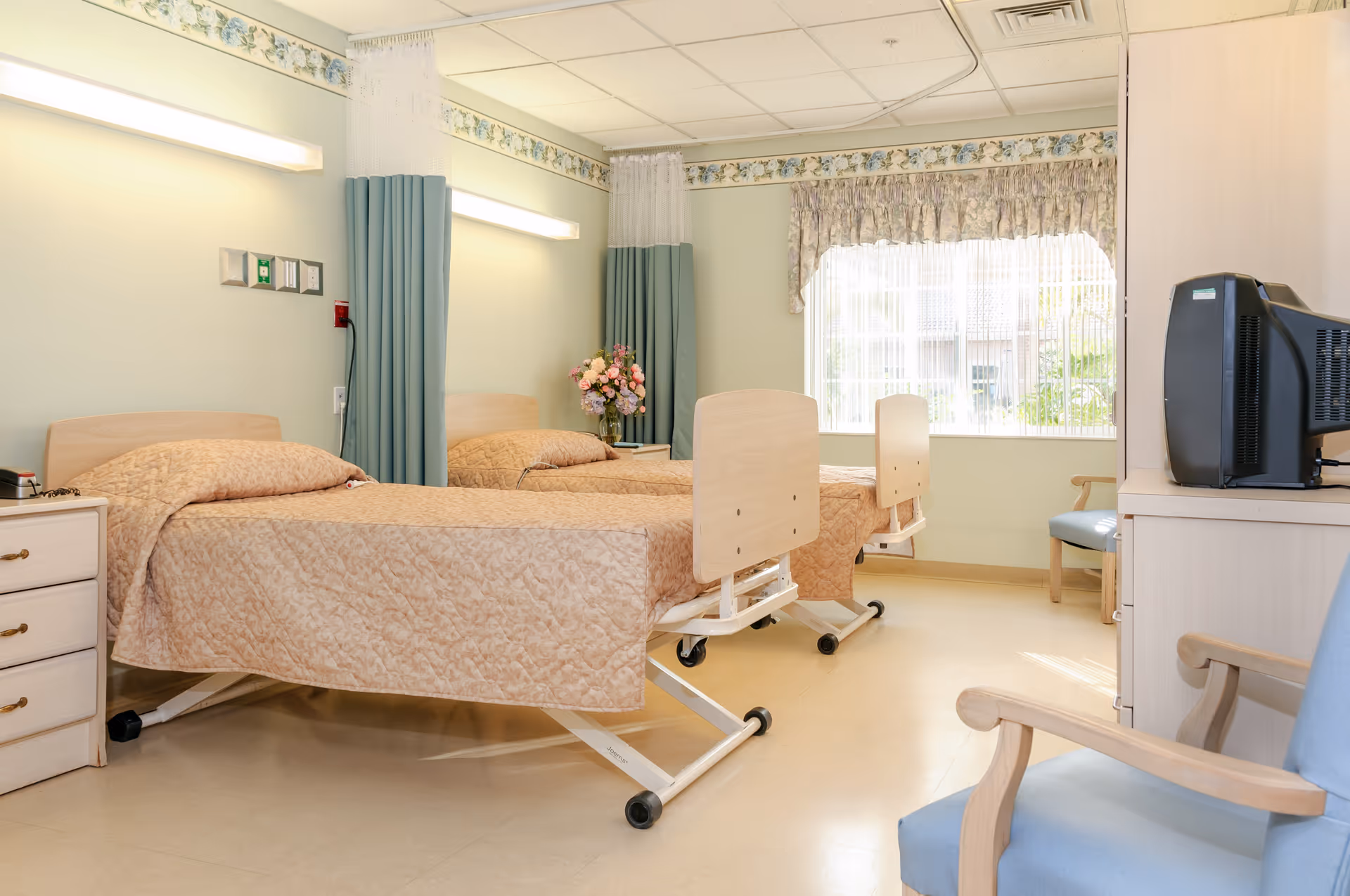 A bright and clean room with two hospital-style beds covered in beige quilts, separated by a green privacy curtain. There is a nightstand with a vase of flowers between the beds, a window with floral curtains letting in natural light, a TV on a cabinet, and two wooden chairs with blue cushions.