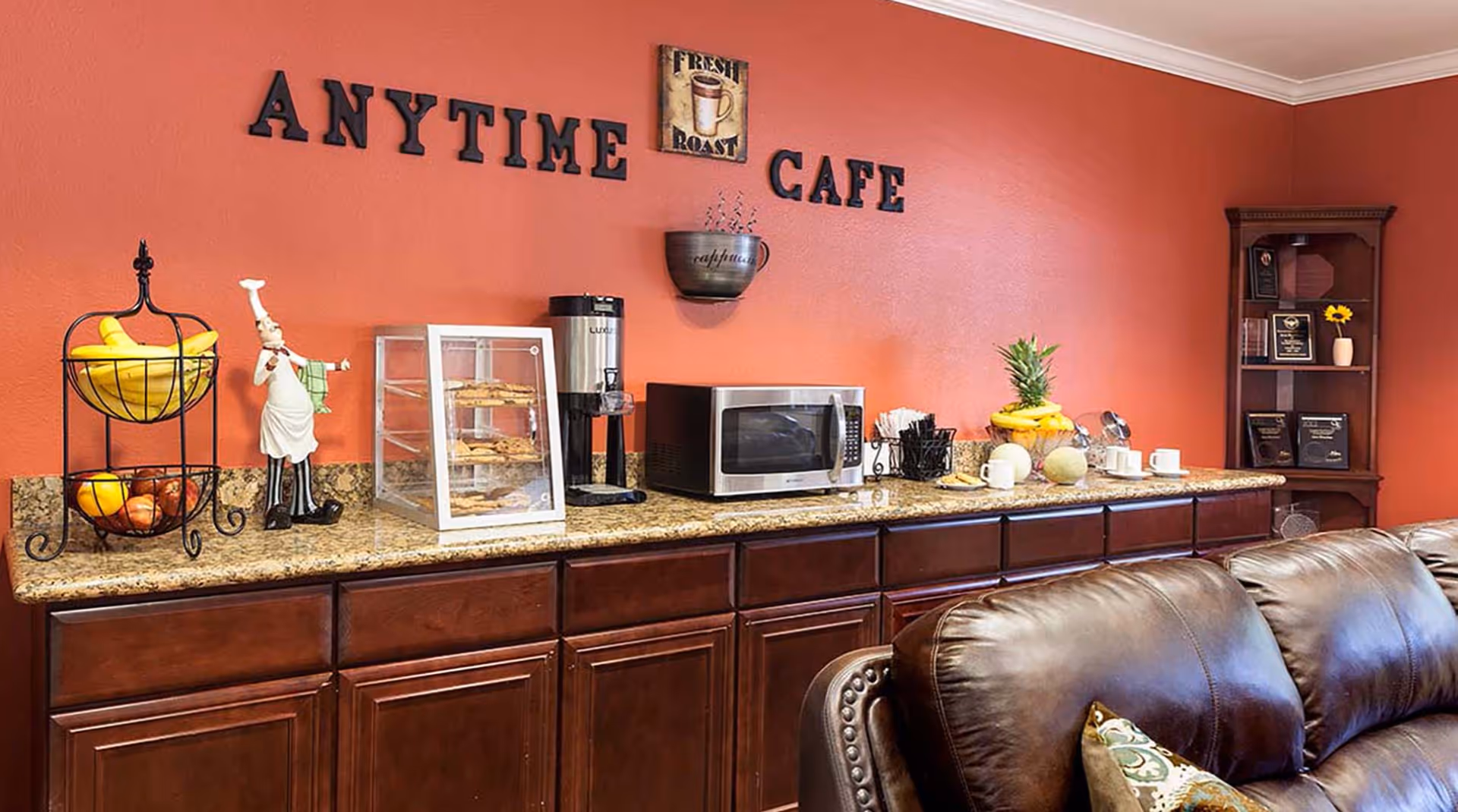 Interior view of a cozy cafe area with a red wall featuring the words 'ANYTIME CAFE'. A countertop holds a tiered fruit basket with bananas and apples, a chef figurine, a display case with pastries, a coffee dispenser, a microwave, and a fruit bowl with a pineapple. There is a dark wooden cabinet with awards and a brown leather couch in the foreground.