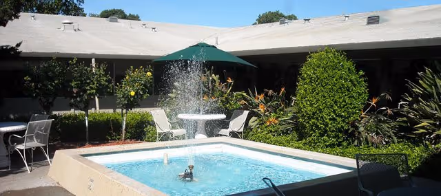 Courtyard with a central fountain pool surrounded by chairs, a green umbrella, and landscaped shrubs in front of a single-story building.