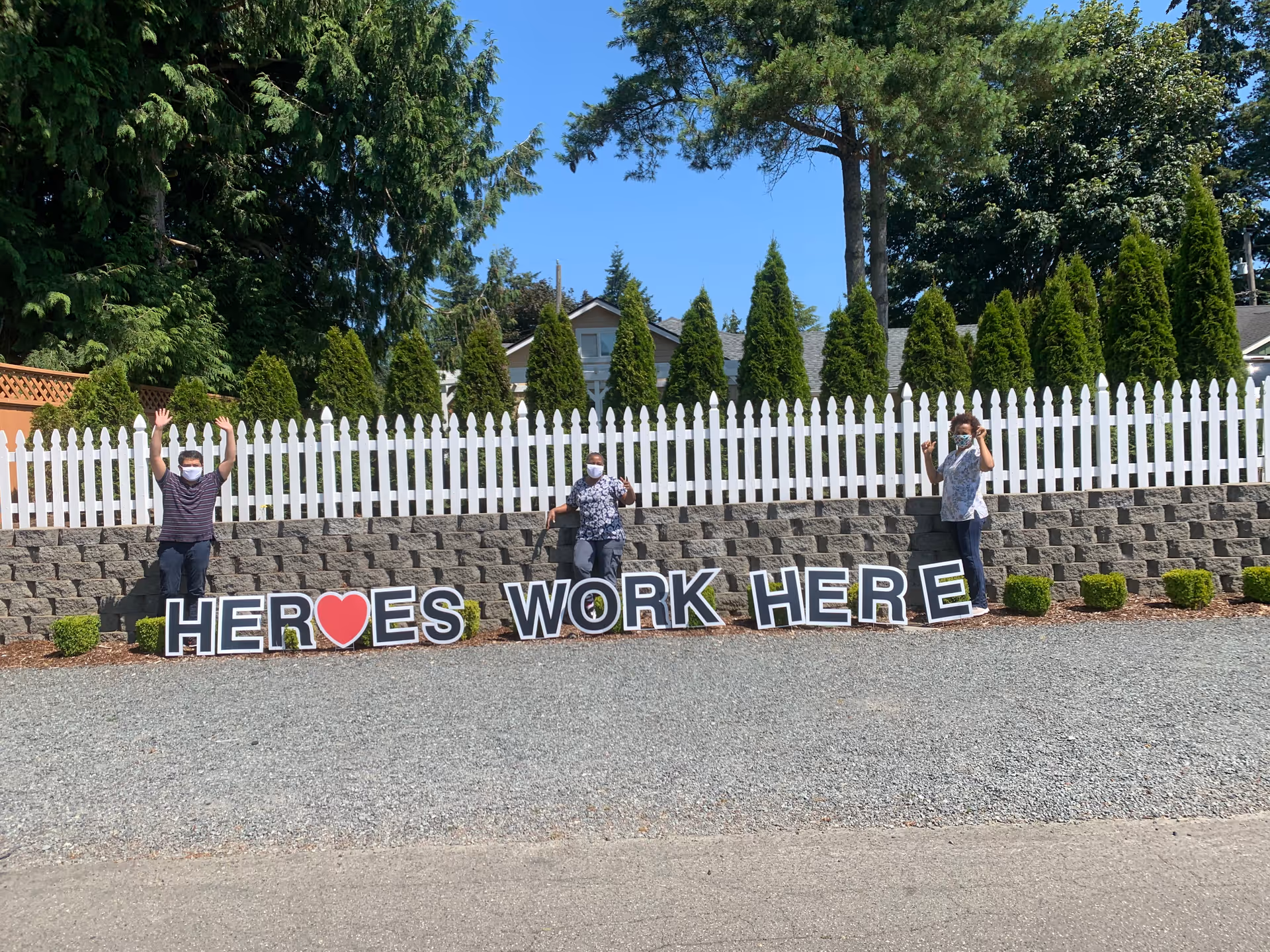 Three people wearing masks standing outdoors in front of a white picket fence and greenery, holding large letters that spell out 'HEROES WORK HERE' with a red heart symbol replacing the letter O in HEROES.