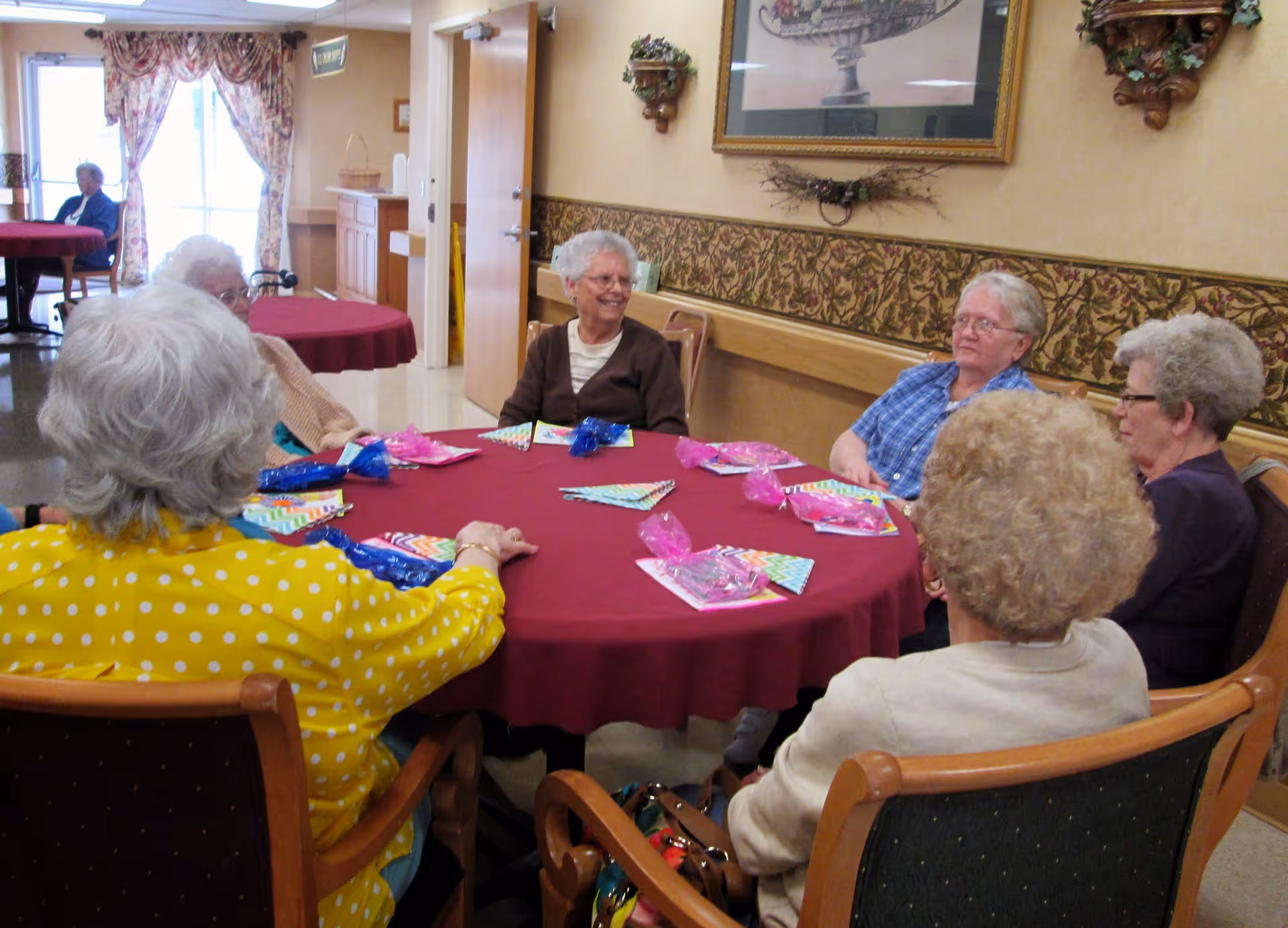 A group of elderly women sitting around a round table covered with a red tablecloth in a common room. The table has colorful napkins and small wrapped gift bags. The room has patterned wallpaper, a framed picture on the wall, and floral curtains on the windows.
