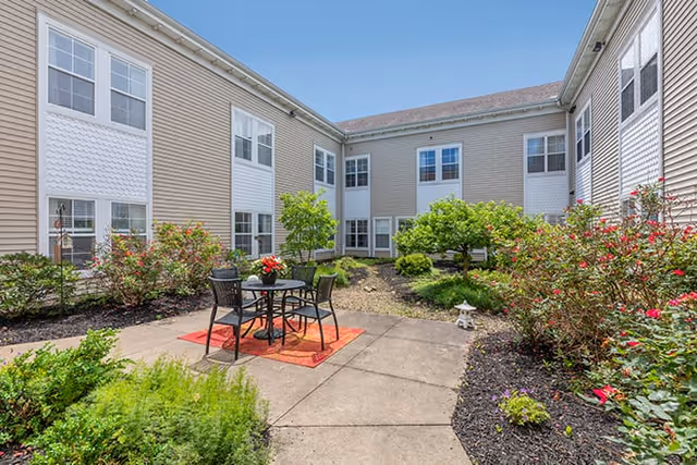 Outdoor courtyard area at Brookdale Mentor with a small round table and four chairs on a red outdoor rug, surrounded by green shrubs, flowering plants, and trees. The courtyard is enclosed by a two-story beige building with multiple windows under a clear blue sky.