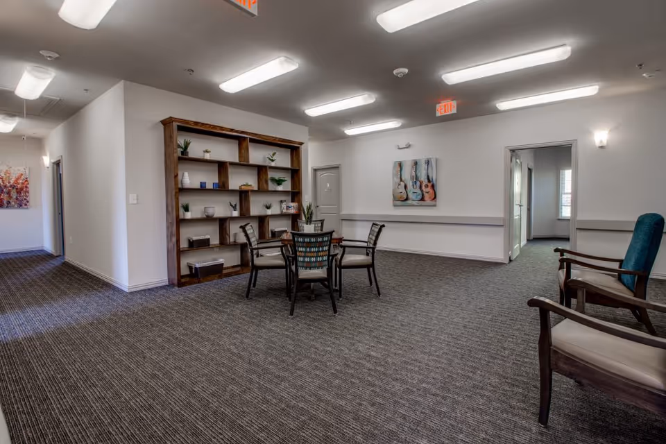 Spacious assisted-living common room with a round table and chairs, a large bookshelf, and armchairs.