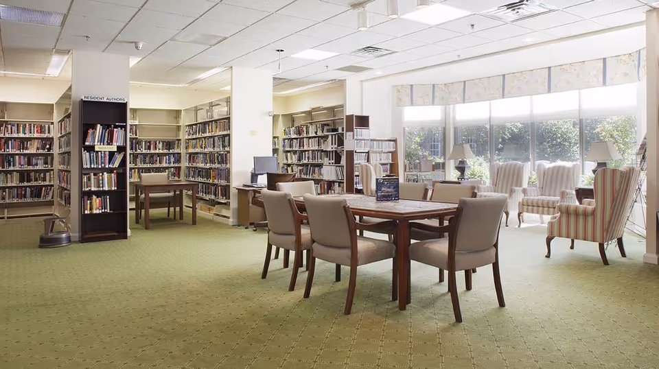 Well-lit library/reading room with bookshelves, a central table with chairs, and upholstered armchairs by large windows.