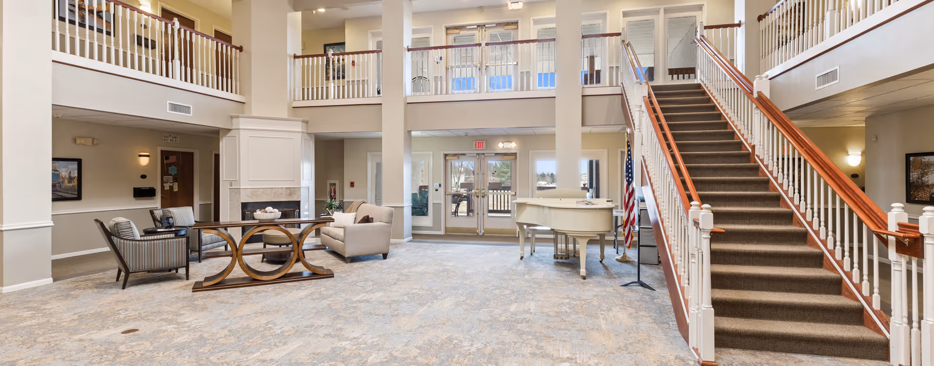 Spacious senior living facility common area with high ceilings, a staircase with wooden handrails on the right, a white grand piano near the entrance doors, and seating arrangements including chairs and a sofa around a wooden table. The area is well-lit with natural light coming through glass doors and windows.