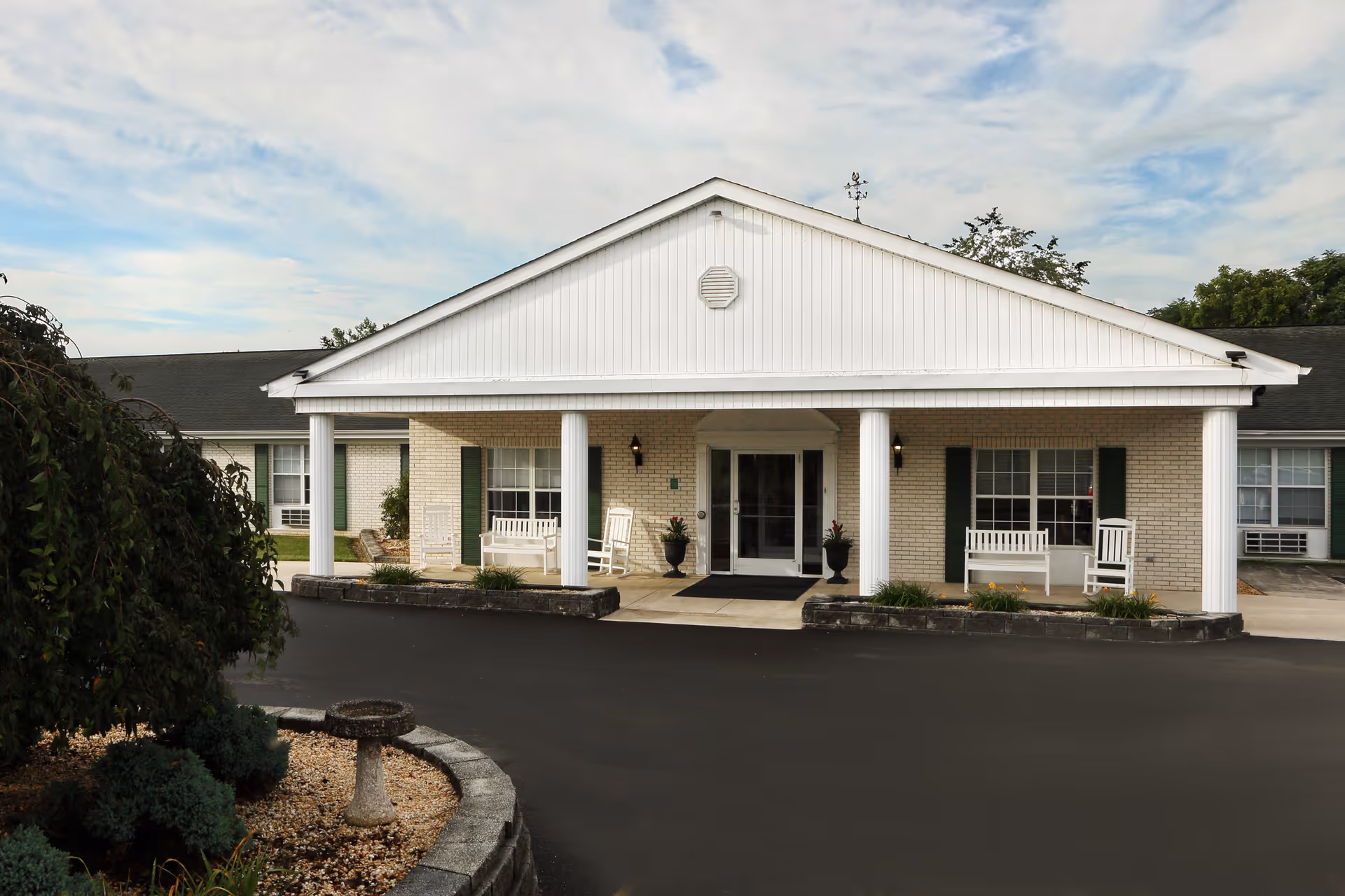 Front exterior of a single-story building with a covered entrance supported by white columns, benches on the porch, and a paved driveway.