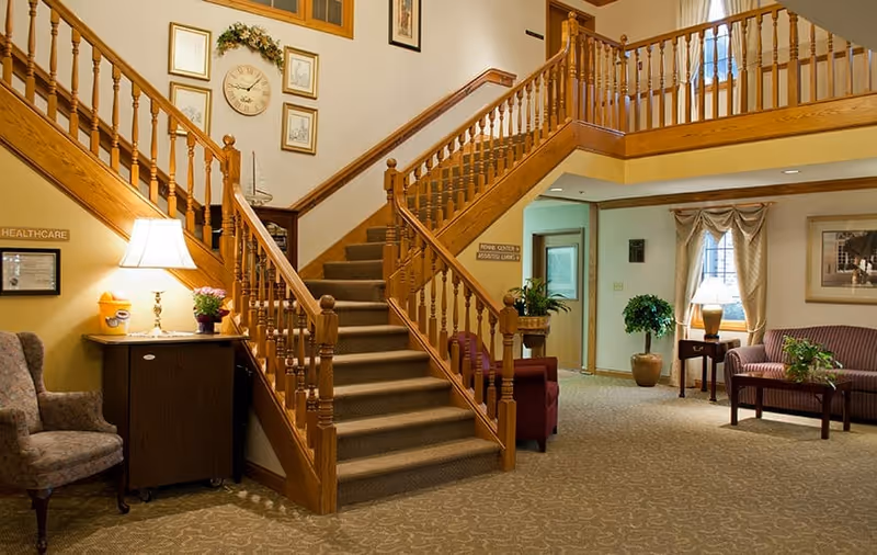Interior view of a senior living facility lobby with a central carpeted staircase featuring wooden railings. The area includes comfortable seating with armchairs and a sofa, side tables with lamps, potted plants, framed pictures on the walls, and a warm, inviting ambiance.