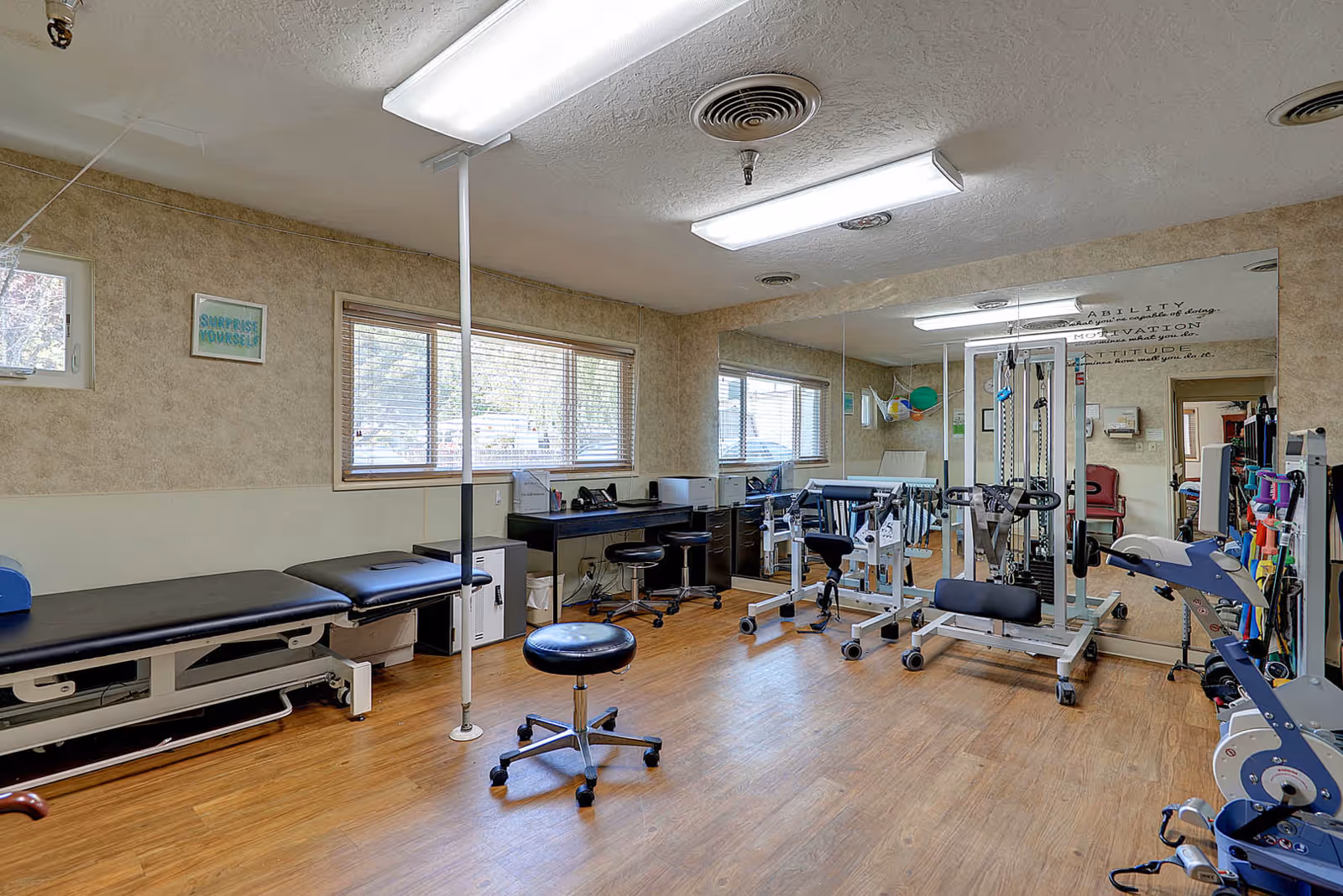 A physical therapy or rehabilitation room with exercise equipment, a treatment table, stools, a desk with office supplies, and large mirrors on one wall. The room has wood flooring, beige walls, and windows with blinds letting in natural light.