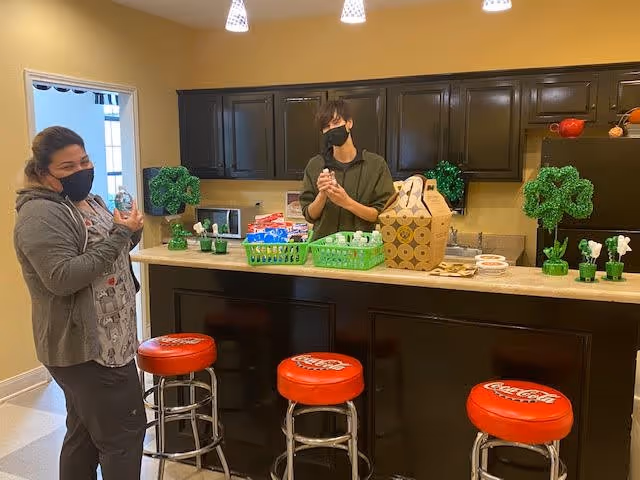 Two people wearing face masks standing behind a kitchen counter with three red Coca-Cola bar stools in front. The counter has green baskets with various items, a cardboard carrier, and small green shamrock decorations. The background shows dark brown cabinets and a microwave.