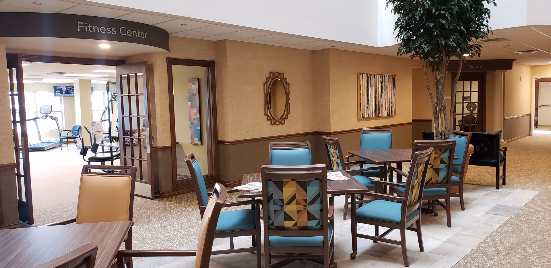 Interior view of a common area in a senior living facility with several tables and chairs arranged for seating. To the left, there is an open doorway leading to a fitness center with exercise equipment visible. On the right side, there is a hallway with a sign for a salon. The area is well-lit with natural light and decorated with wall art and a large indoor plant.