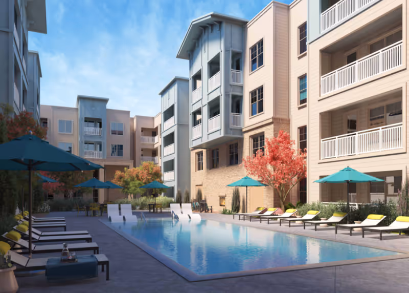 Outdoor swimming pool area at a senior living facility with lounge chairs and umbrellas around the pool, surrounded by multi-story residential buildings and trees with autumn-colored leaves under a blue sky.
