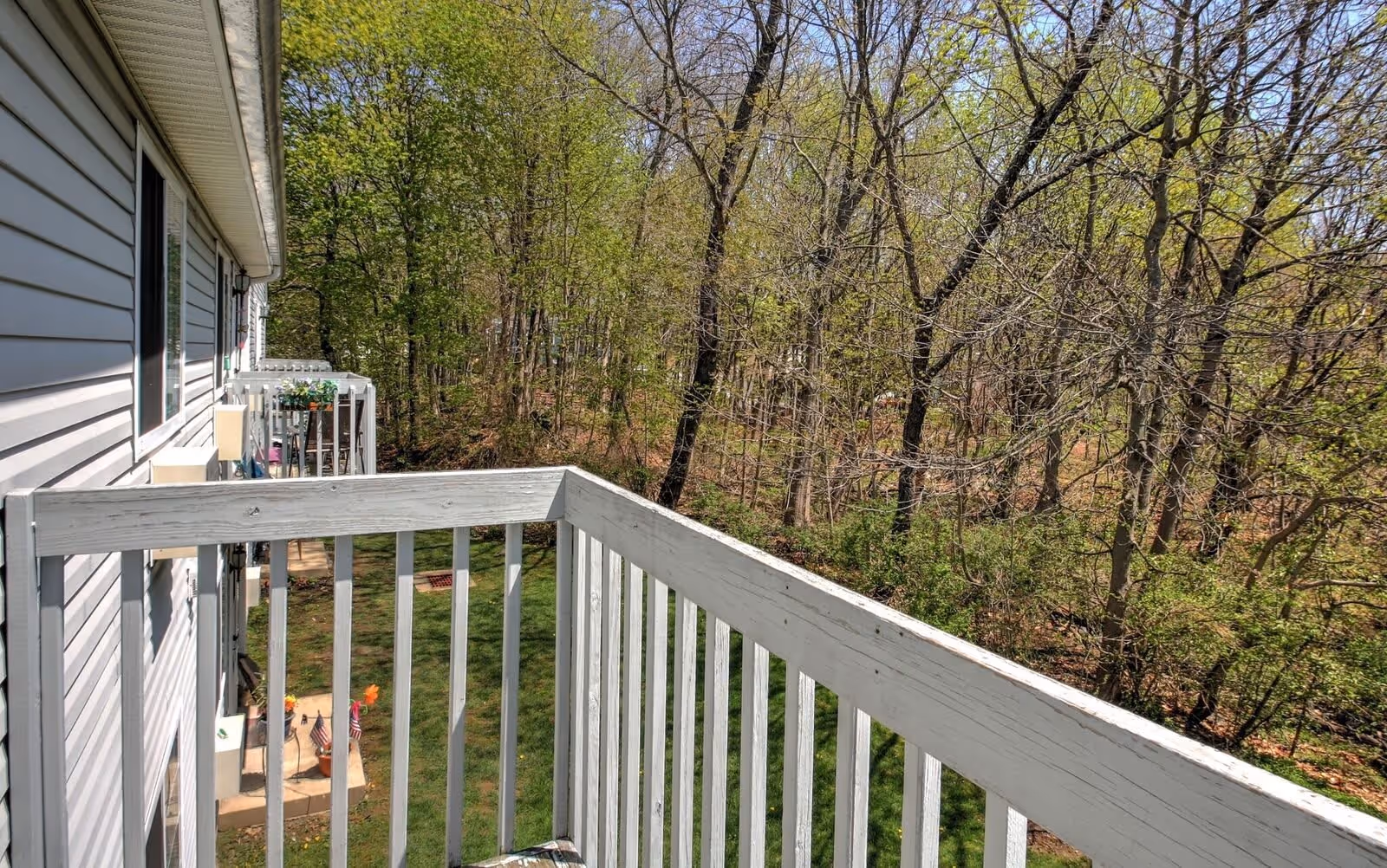 View from a white wooden balcony overlooking a grassy area and a wooded area with trees. The side of a building with white siding and several balconies is visible on the left side of the image.