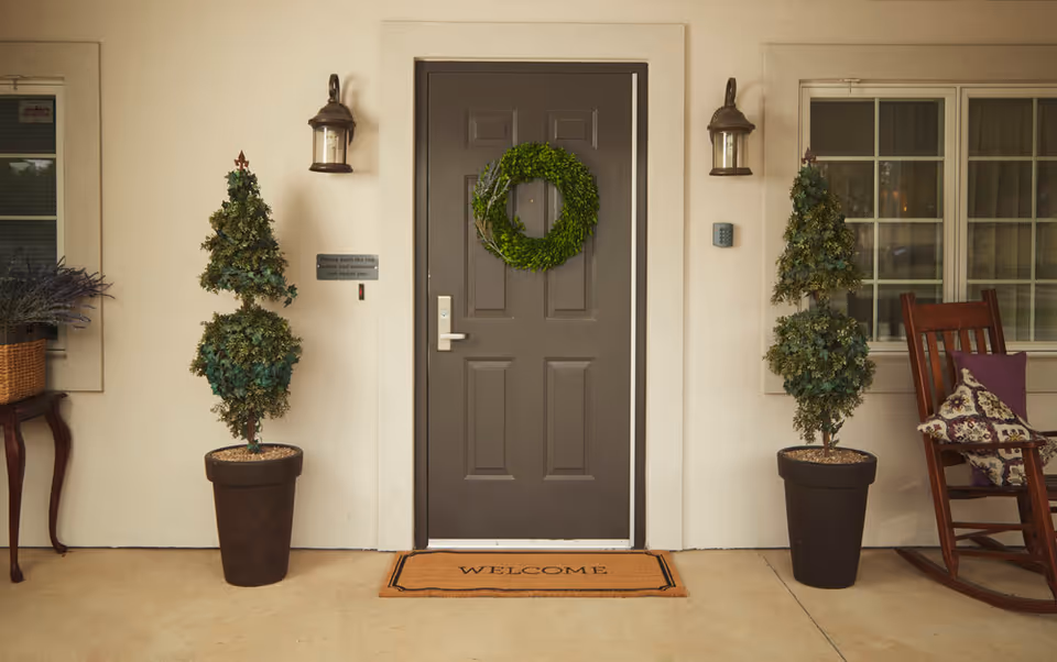 Front entrance with a gray door decorated with a green wreath, a 'WELCOME' doormat, two potted topiaries, and a rocking chair on the porch.