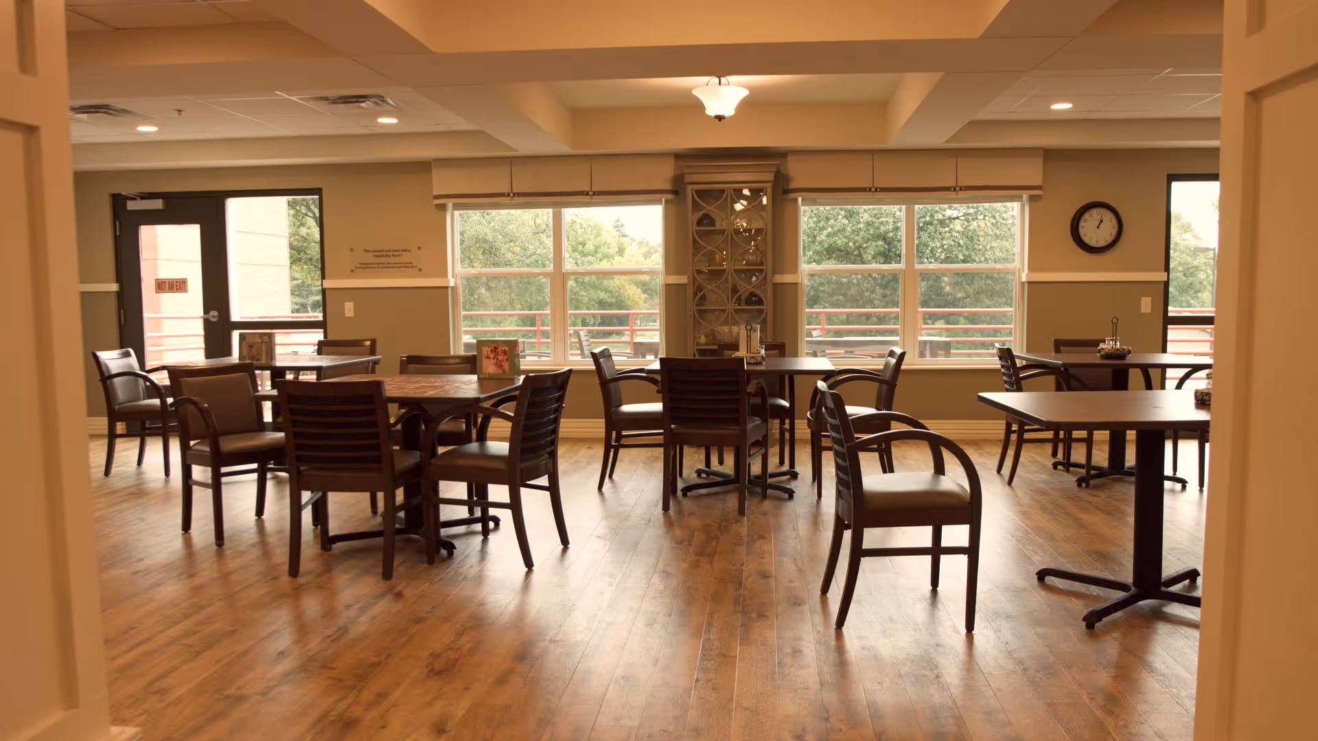 A dining room with several wooden tables and chairs arranged on a wooden floor. Large windows let in natural light and show greenery outside. A clock is mounted on the wall, and there is a door on the left side of the room.
