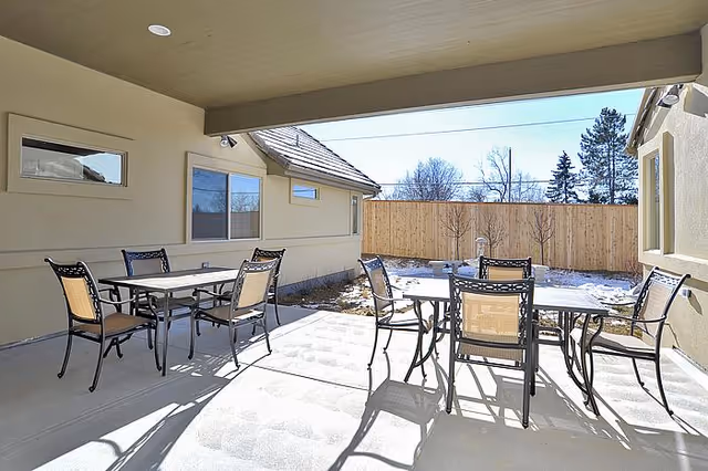 Outdoor covered patio area with two metal tables and eight chairs on a concrete floor, adjacent to a beige building with windows, and a wooden privacy fence enclosing a yard with some snow and leafless trees.