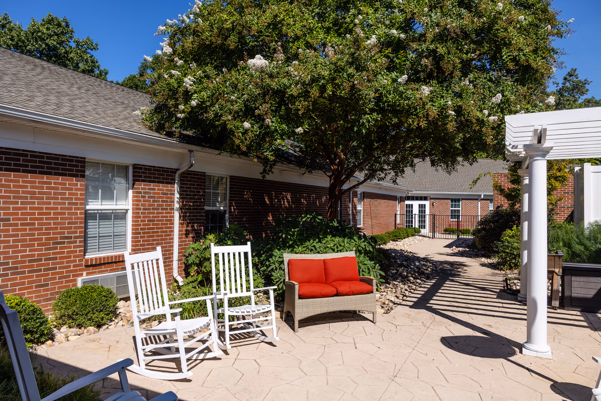 Sunny courtyard with white rocking chairs and a red-cushioned loveseat beside a brick building, a tree and a pergola.