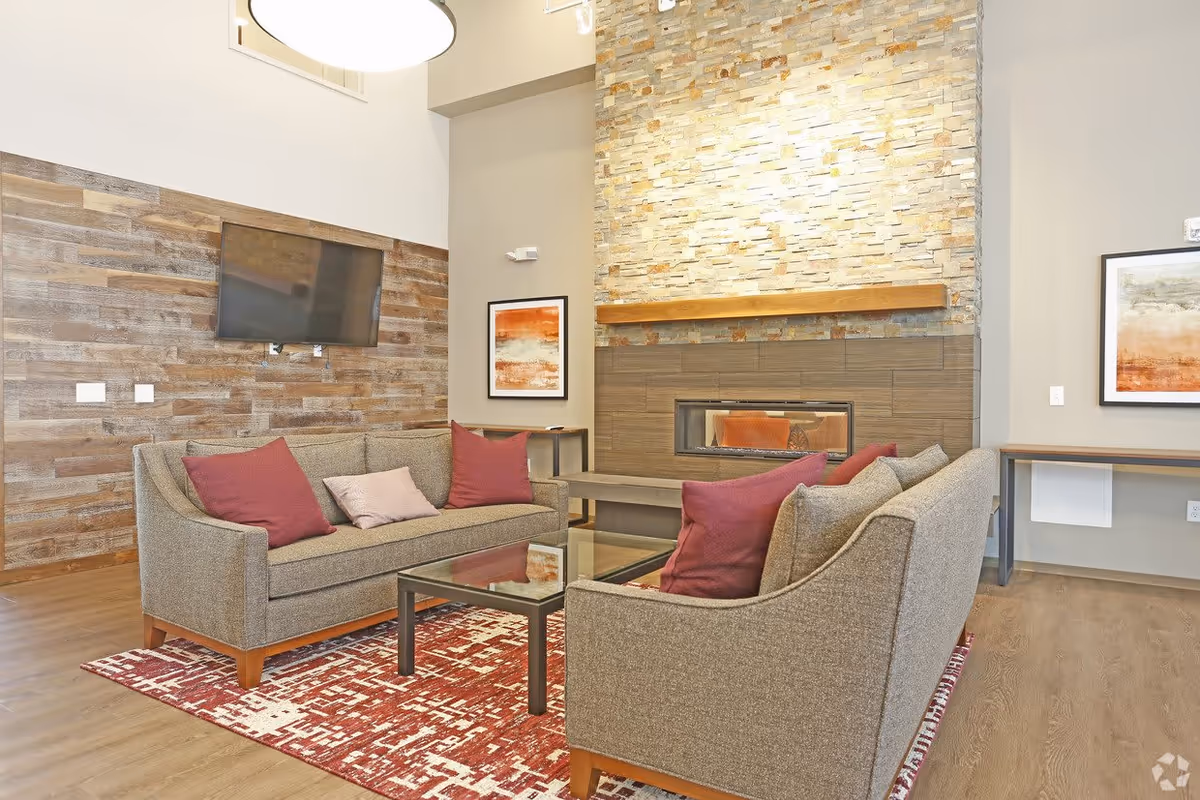 A cozy living room area with two gray sofas adorned with red and pink pillows, a glass coffee table on a red patterned rug, a wall-mounted flat-screen TV, and a modern fireplace with a stone and wood accent wall. The room has wooden flooring and neutral-colored walls with framed artwork.