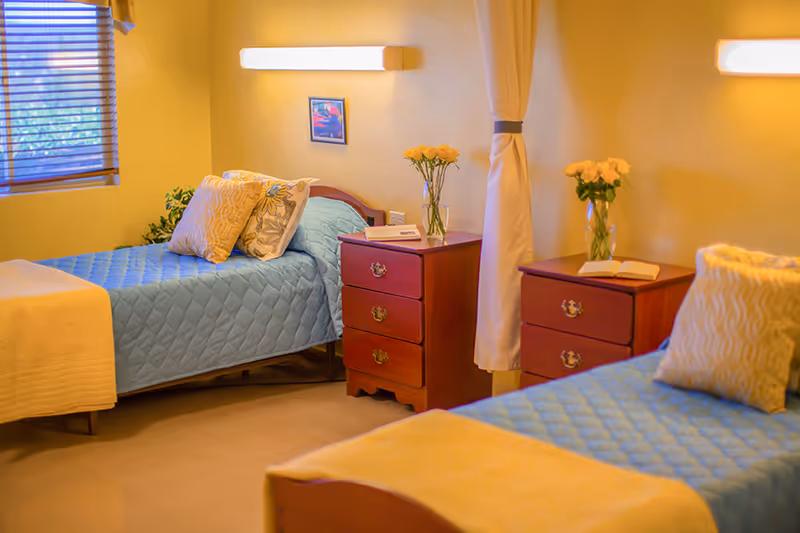 A cozy room with two single beds covered in blue quilted bedspreads and beige blankets. Each bed has decorative pillows. Between the beds are two wooden nightstands with drawers, each holding a vase with yellow flowers. The room has warm yellow walls, a window with blinds, and soft wall-mounted lighting above each bed.