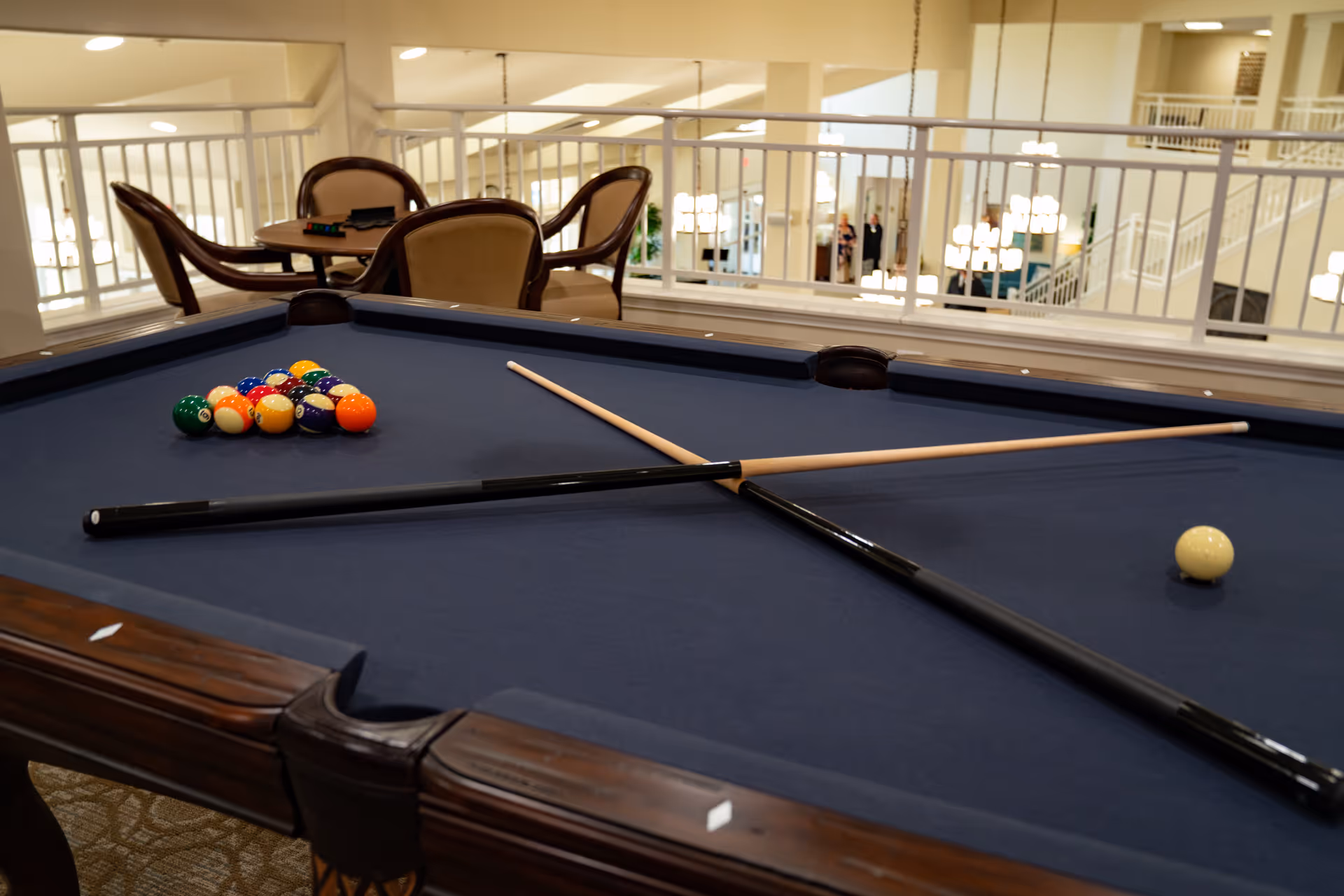 A pool table with a blue felt surface set up for a game with a triangle of billiard balls and two crossed pool cues resting on the table. In the background, there are chairs and a table, with a railing overlooking a well-lit interior space with chandeliers and stairs.