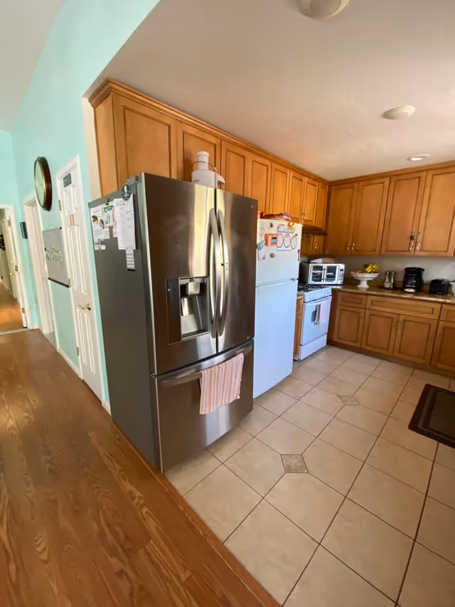 A kitchen with wooden cabinets, a stainless steel refrigerator with a towel hanging on the handle, a white refrigerator with magnets, a stove, a microwave, a coffee maker, and a fruit bowl on the counter. The floor is tiled and the adjacent hallway has wooden flooring.