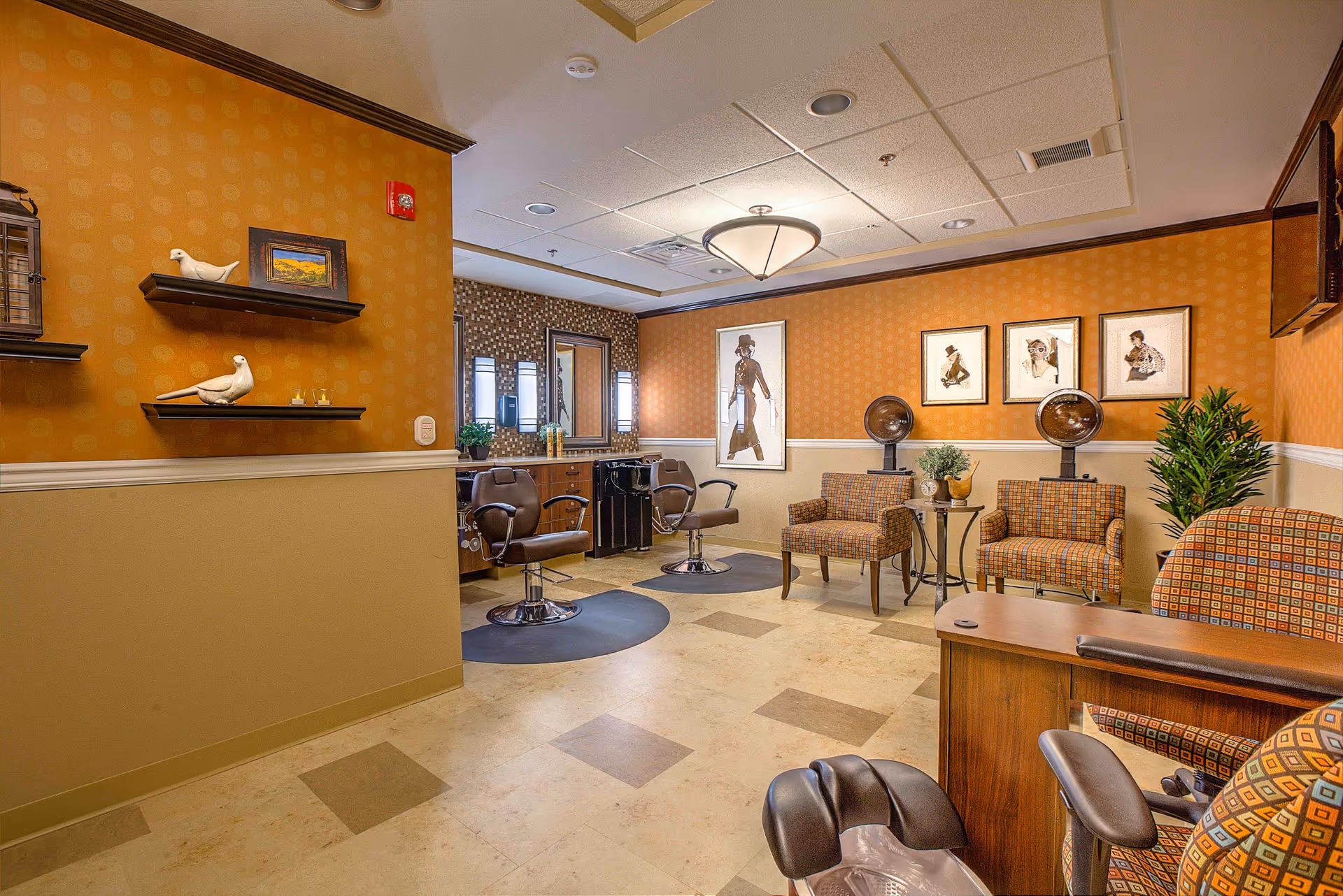 Interior view of a salon area in an assisted living facility featuring two salon chairs with mirrors and hair dryers, a seating area with patterned armchairs and a small table, decorative wall art, and warm-toned walls with shelves displaying small decorative items.