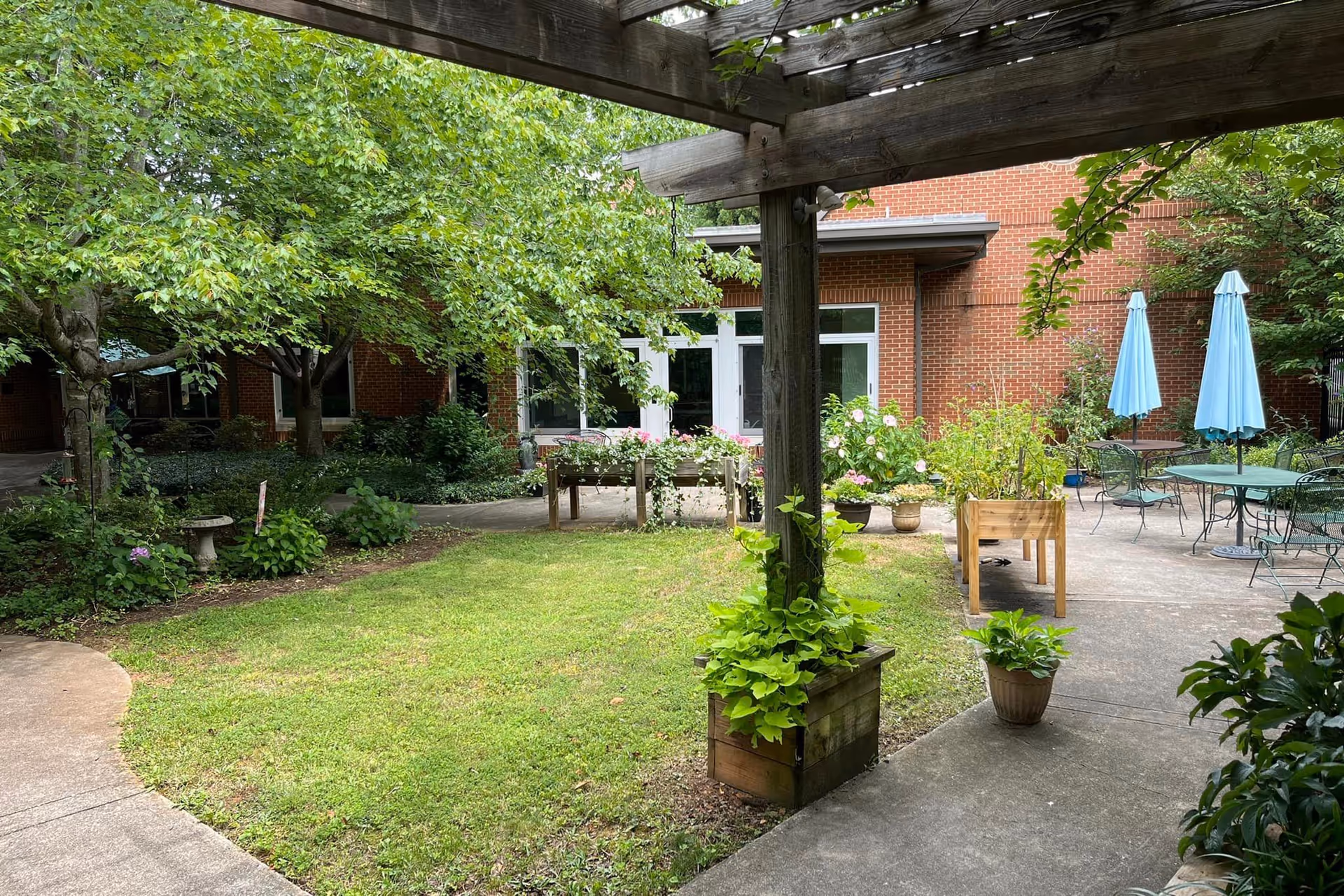 A peaceful outdoor courtyard area with green grass, trees, and various potted plants. There is a wooden pergola with climbing plants, a concrete pathway, and several tables with blue umbrellas. The background shows a brick building with white-framed windows and doors.