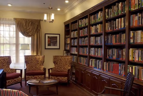 A cozy library room with a large wooden bookshelf filled with books along one wall. There are three patterned armchairs with red cushions arranged around a small round wooden coffee table. A window with sheer curtains and drapes lets in natural light, and a floor lamp stands beside the chairs. A framed picture hangs on the wall above the chairs.
