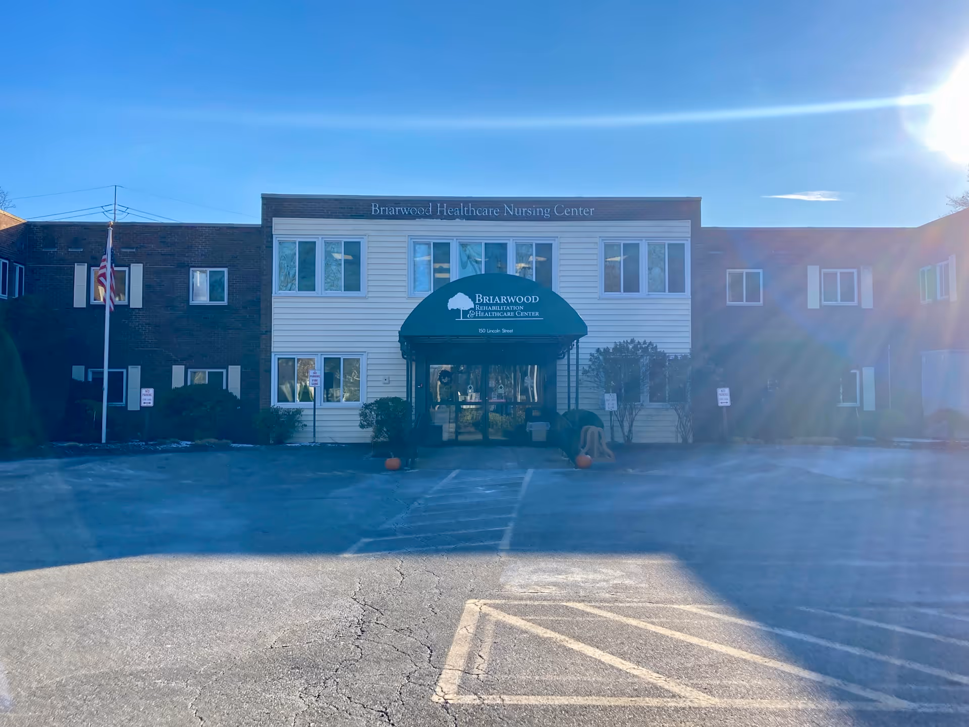 Front exterior view of Briarwood Rehabilitation & Healthcare Center building with a green awning over the entrance, an American flag on a pole to the left, and a clear blue sky with sunlight creating lens flare.