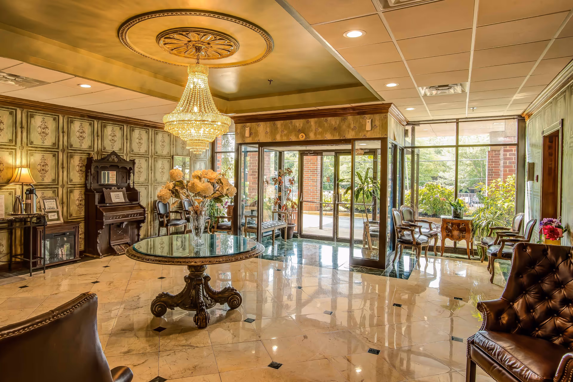 A spacious and elegant lobby area with a large ornate chandelier hanging from the ceiling. The floor is polished marble with black diamond accents. There is a round glass table with a decorative base in the center, holding a vase of flowers. To the left, there is an antique-style piano and a small table with a lamp and framed signs. The walls have a vintage patterned wallpaper. The entrance features glass doors with large windows allowing natural light to fill the space. Several classic-style chairs and small tables are arranged near the windows and along the walls.