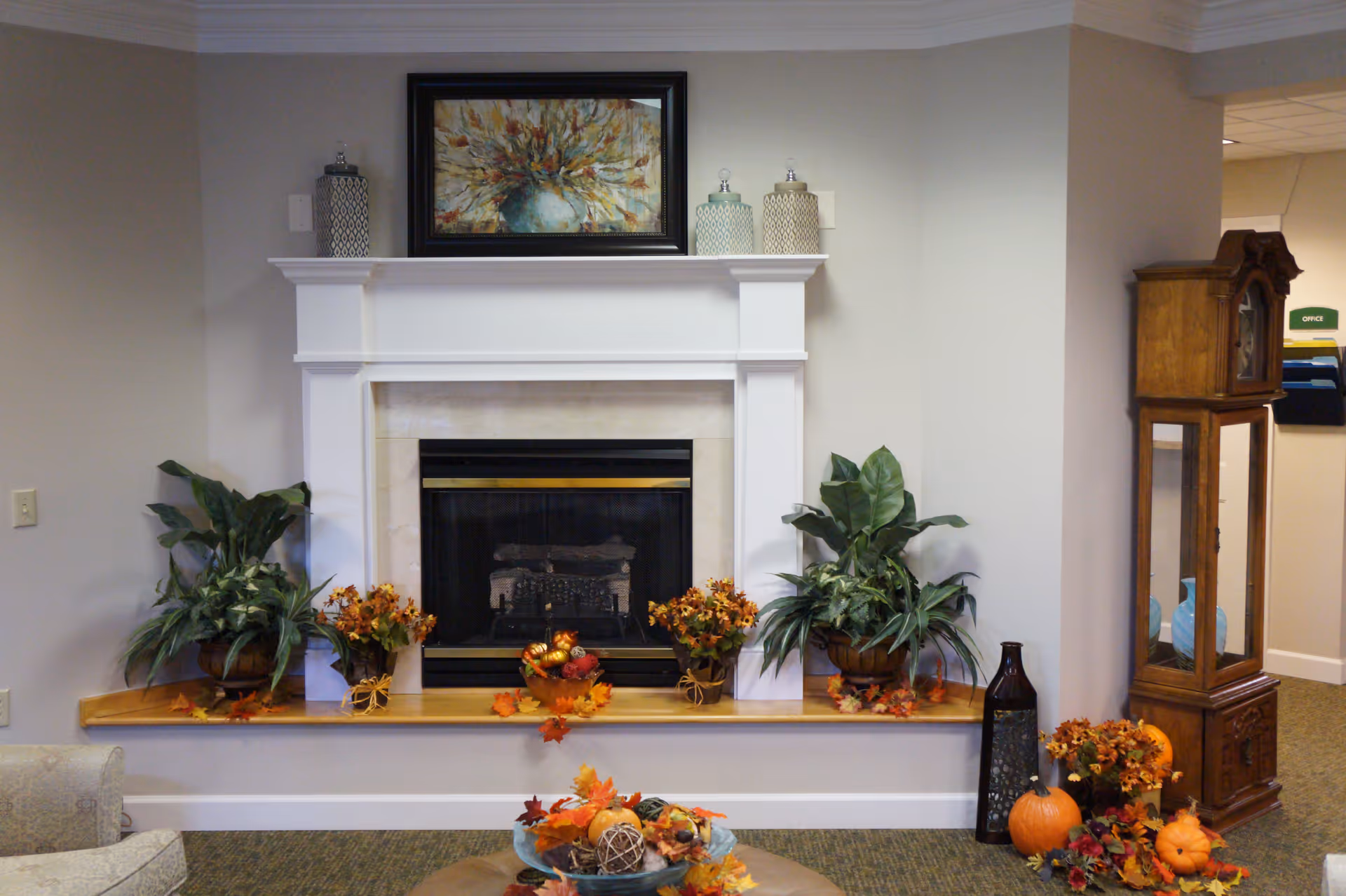 A cozy indoor living area featuring a white fireplace with a black and gold insert. The mantel is decorated with a framed floral painting and three decorative jars. On the hearth, there are two potted green plants and autumn-themed floral arrangements. To the right of the fireplace, there is a tall wooden grandfather clock and a collection of pumpkins and fall decorations on the floor. The room has neutral-colored walls and carpeted flooring.