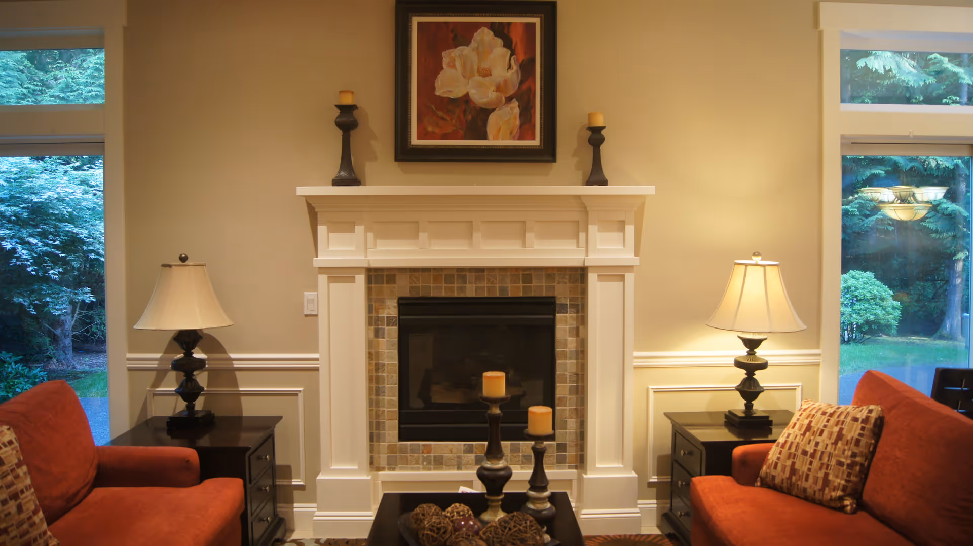 A cozy living room featuring a white fireplace with a tiled surround, flanked by two dark wooden side tables each with a lamp. Two red upholstered armchairs with patterned cushions face each other across a small coffee table with decorative items. Large windows on either side of the fireplace show a green outdoor garden.
