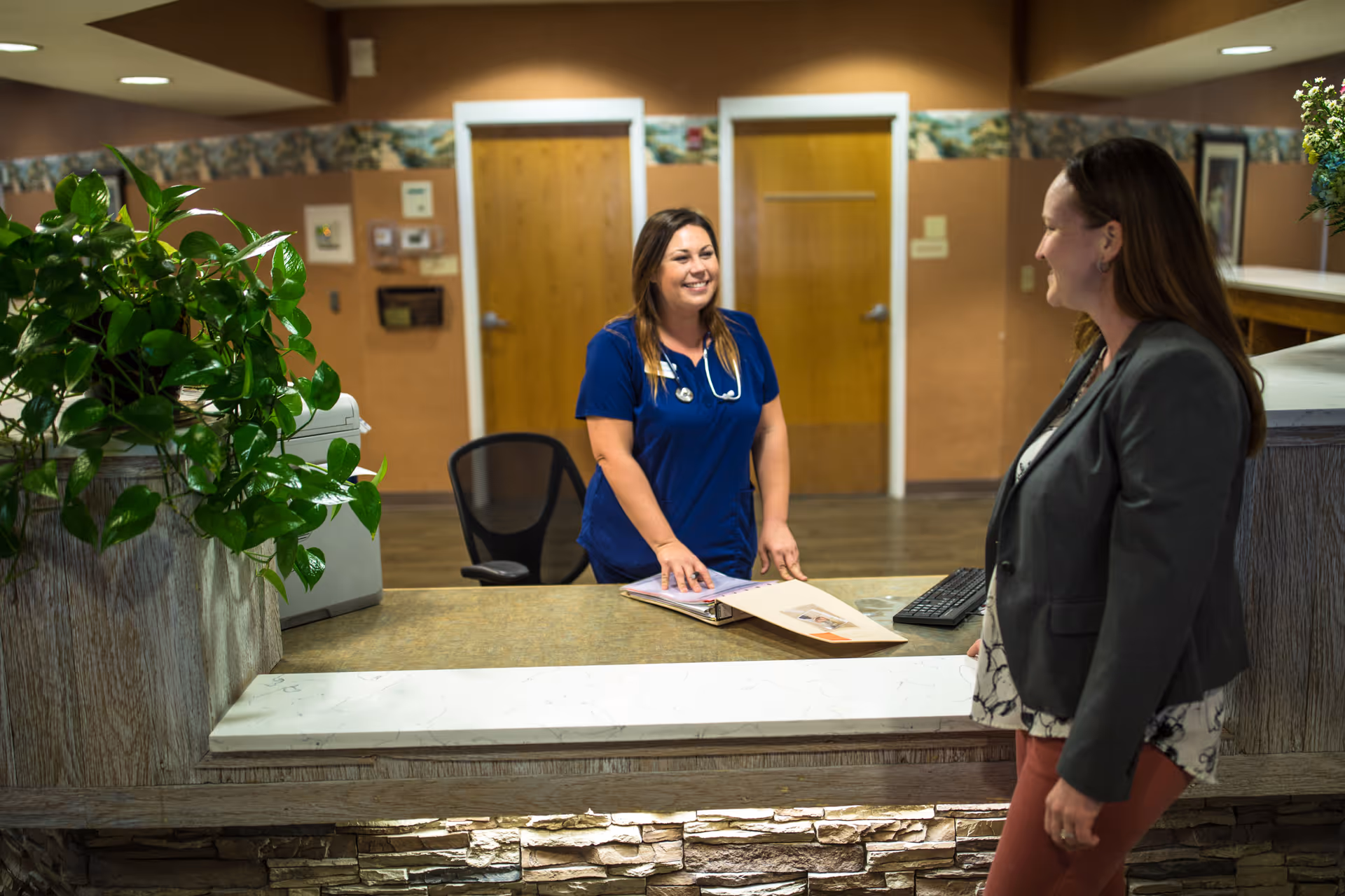 A healthcare worker in blue scrubs stands behind a reception desk smiling and talking to a woman in a gray blazer and patterned blouse. The reception area has a plant on the counter, wooden doors in the background, and warm-toned walls with a decorative border near the ceiling.