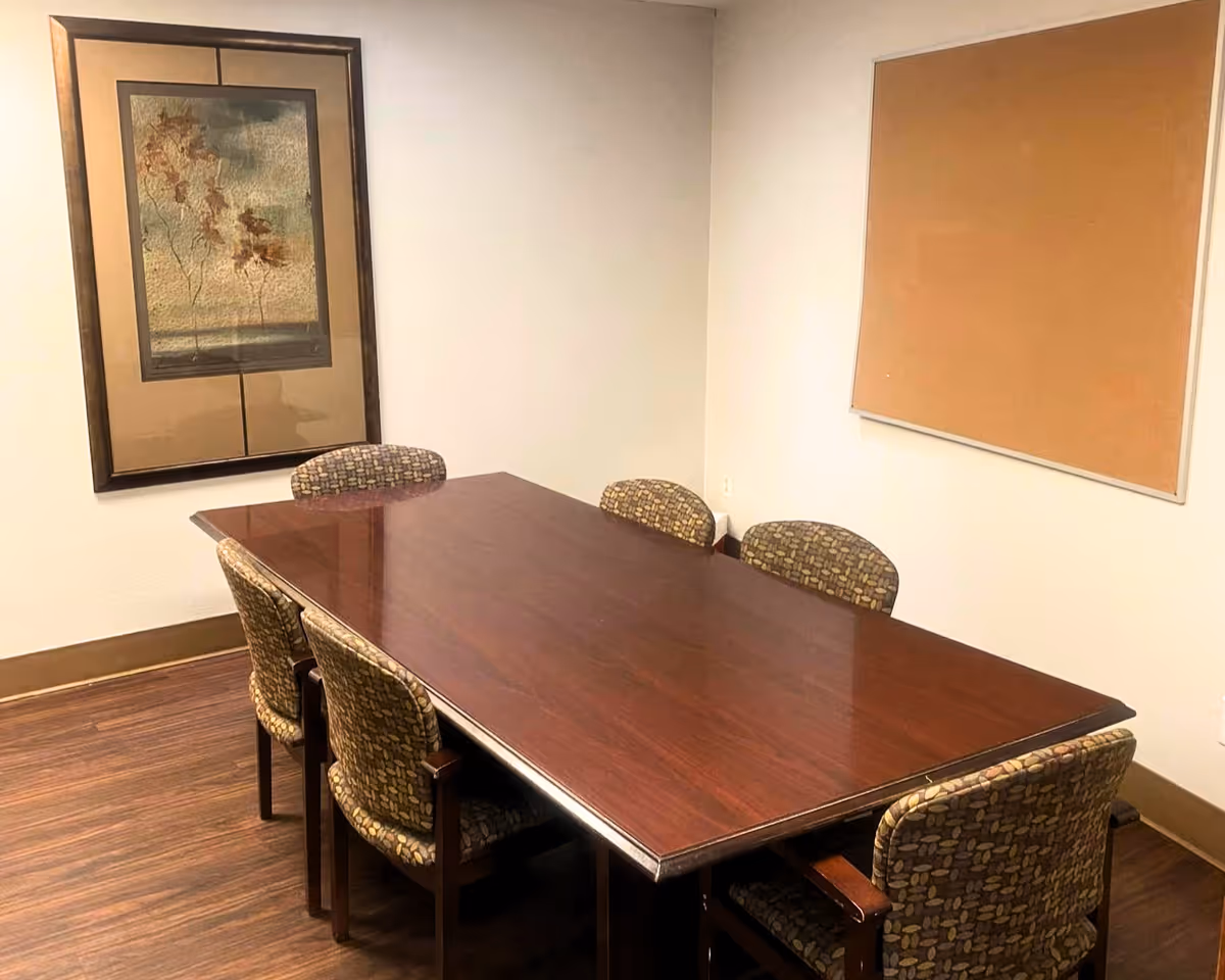 A small conference or meeting room with a rectangular wooden table surrounded by six patterned upholstered chairs. The room has light-colored walls, a framed artwork on one wall, and a large empty cork bulletin board on another wall. The floor is wooden.