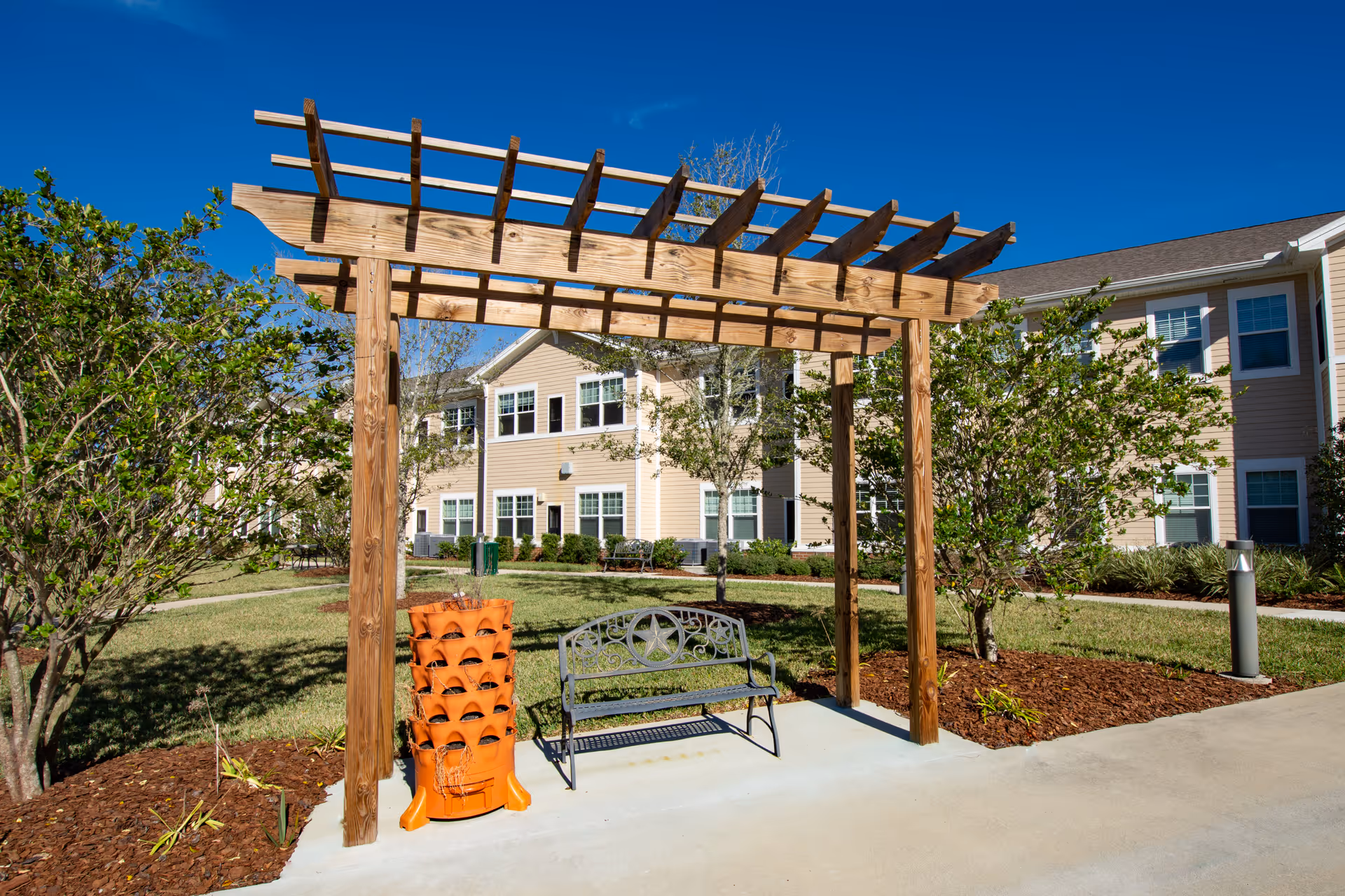 Outdoor seating area at Anthem Lakes featuring a wooden pergola, a metal bench with decorative backrest, and a vertical orange planter. The area is surrounded by green grass, trees, and shrubs with a beige two-story building in the background under a clear blue sky.