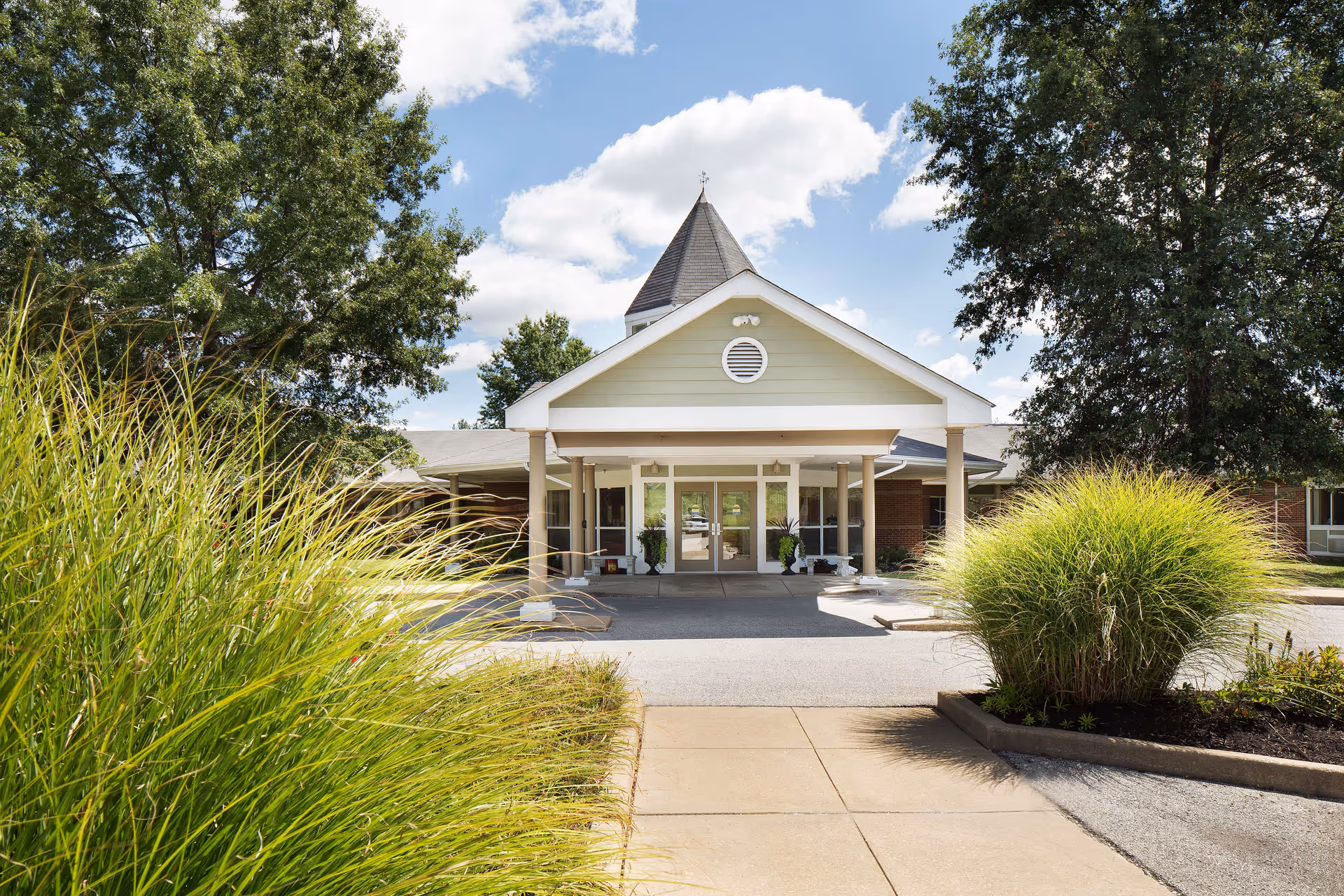 Front entrance of a single-story senior living facility with a covered porte-cochere, potted plants, and landscaped grasses.