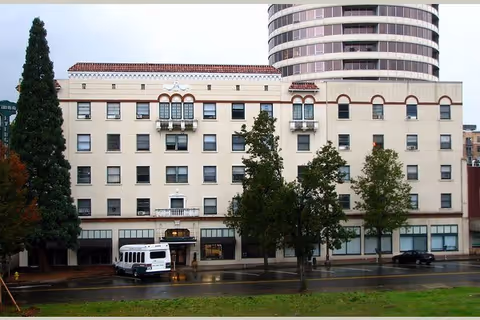 Exterior view of a multi-story senior living facility named The Evergreen Inn with several windows, trees in front, a white shuttle bus parked near the entrance, and a taller round building visible in the background.
