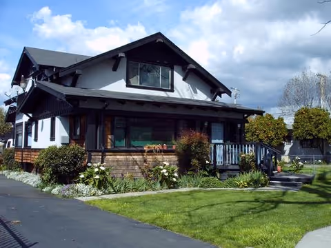 A two-story residential building with a dark roof and white upper walls, featuring a porch with wooden railings and surrounded by green grass, bushes, and a paved driveway under a partly cloudy sky.