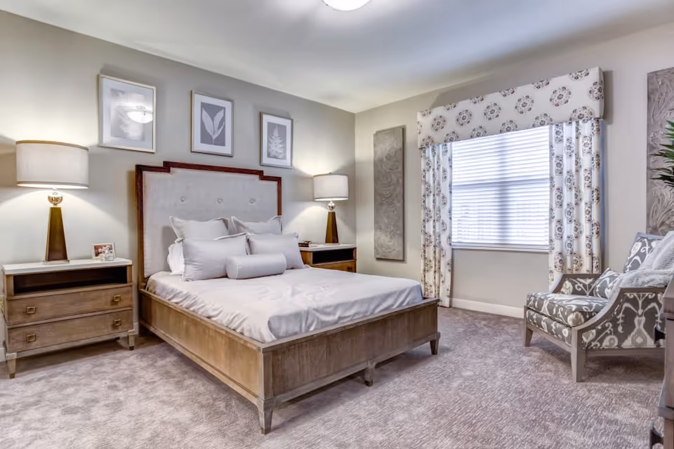 Bright, neutral bedroom with a wooden bed, two bedside tables and lamps, a patterned armchair, and a window with floral curtains.