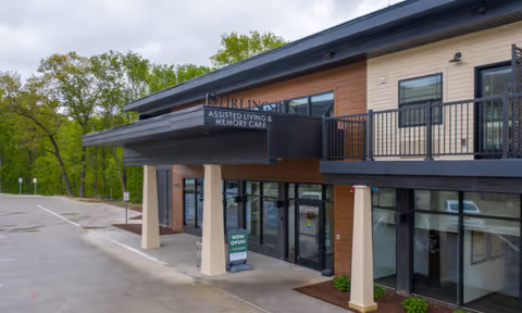 Entrance of Stirlingshire Assisted Living & Memory Care building with a covered drop-off, glass doors, and a 'Now Open' sign.