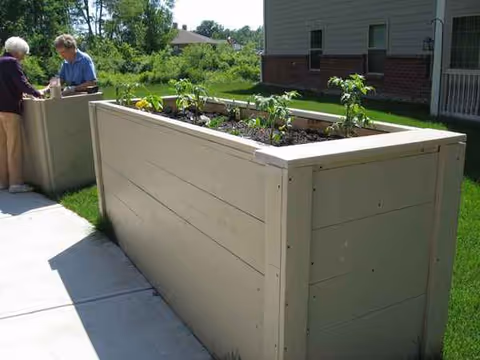 Large raised planter box with plants outside a building, with two people conversing in the background.