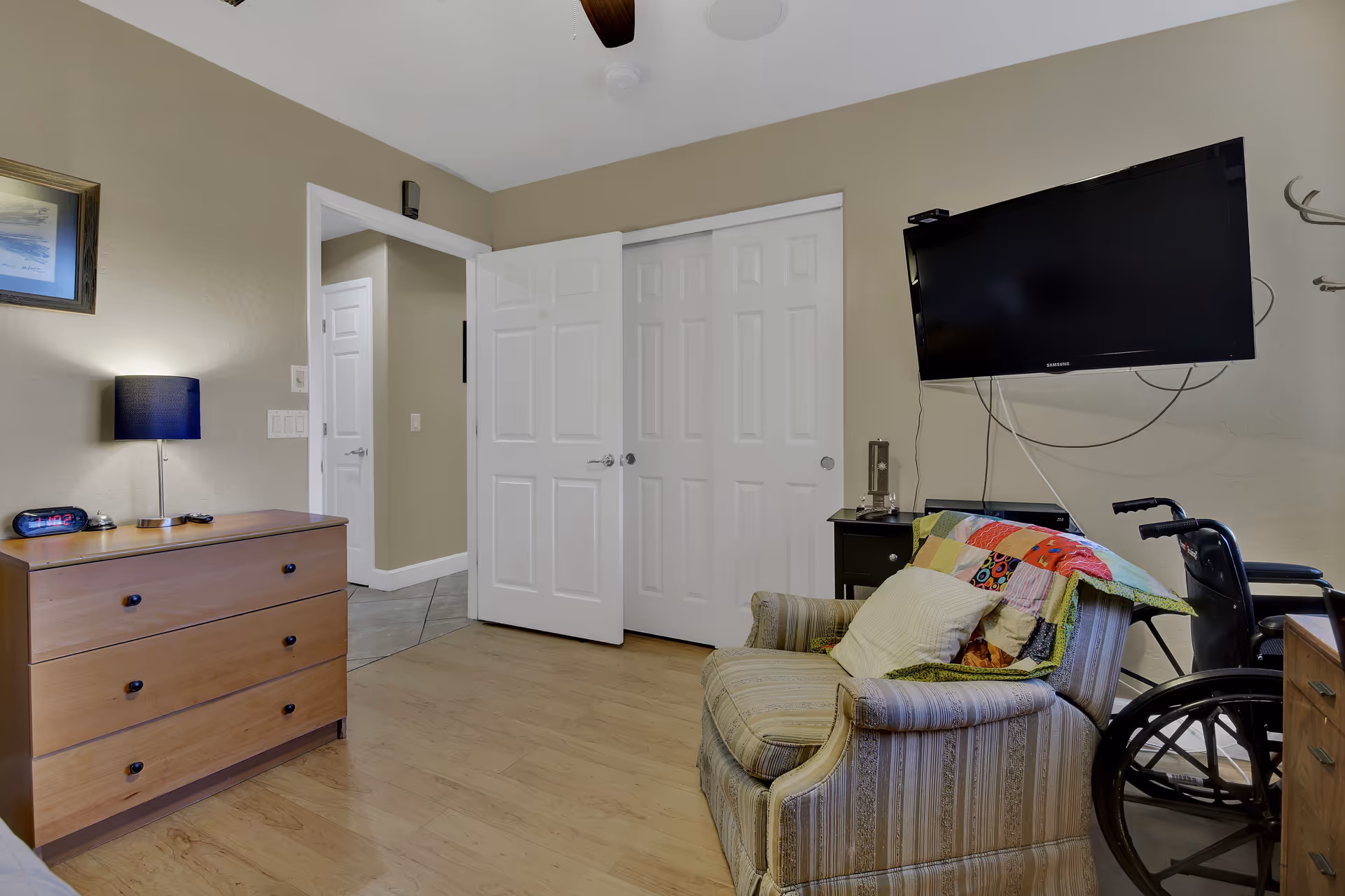 A cozy room with a wooden dresser topped with a lamp and digital clock, a striped armchair with a colorful quilt and pillow, a wheelchair beside the armchair, a wall-mounted flat-screen TV, and white double doors leading to another area.
