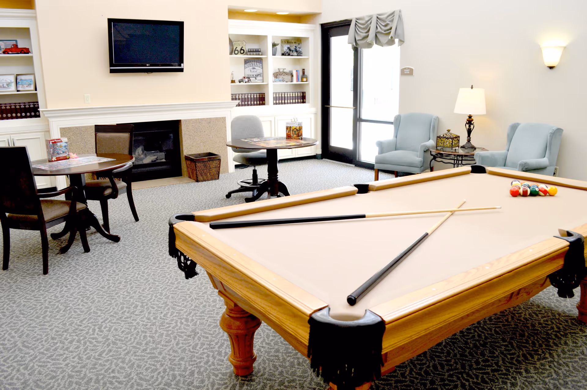 A bright and cozy common area featuring a beige pool table with two cues and a rack of billiard balls, two round tables with chairs, a fireplace with a mounted flat-screen TV above it, built-in shelves with books and decorative items, two light blue armchairs next to a small table with a lamp, and a glass door with a window covered by a valance.