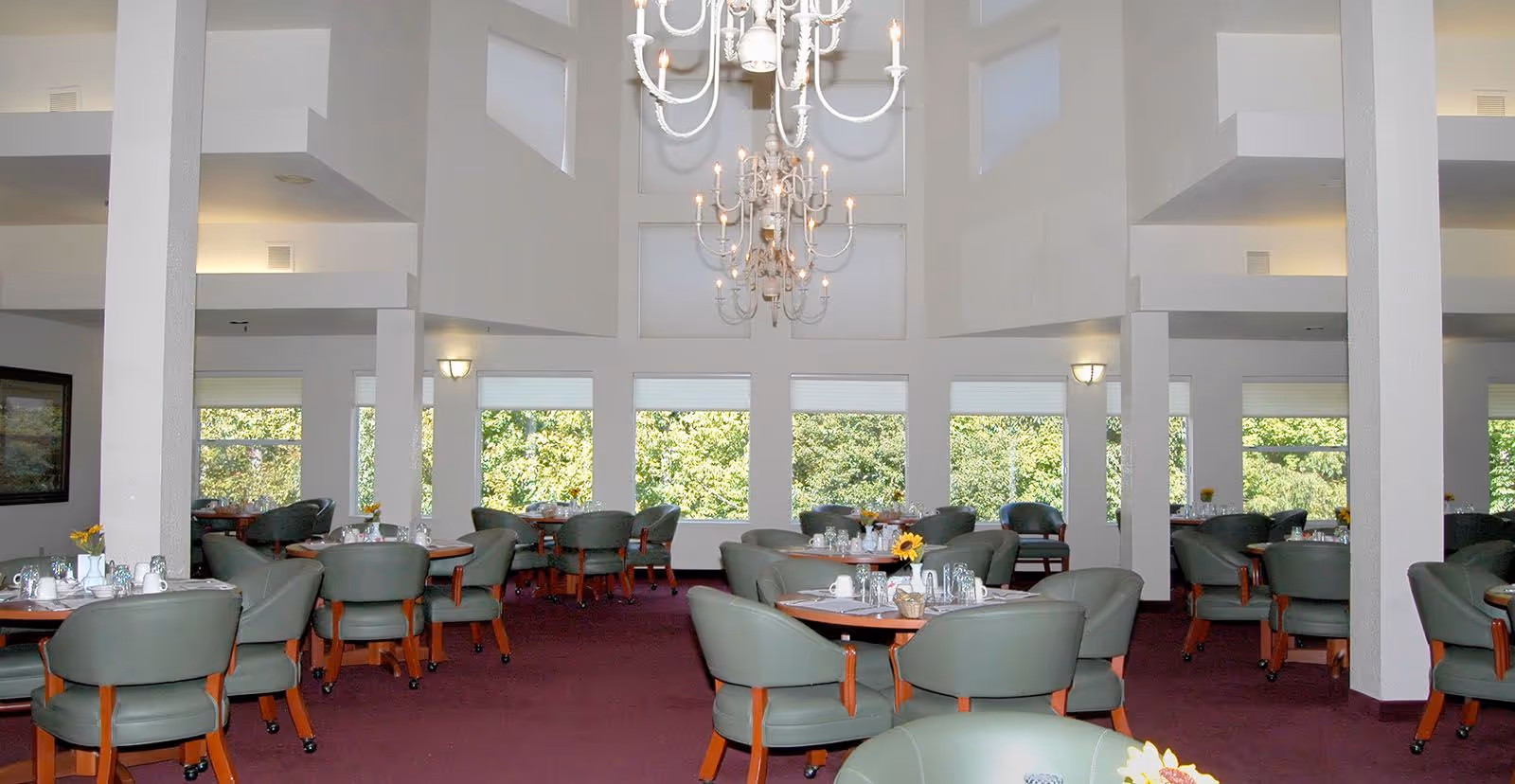 A spacious dining room with multiple round tables surrounded by green cushioned chairs. The room has large windows letting in natural light and a high ceiling with two elegant chandeliers. The carpet is a deep red color and the walls are painted white.