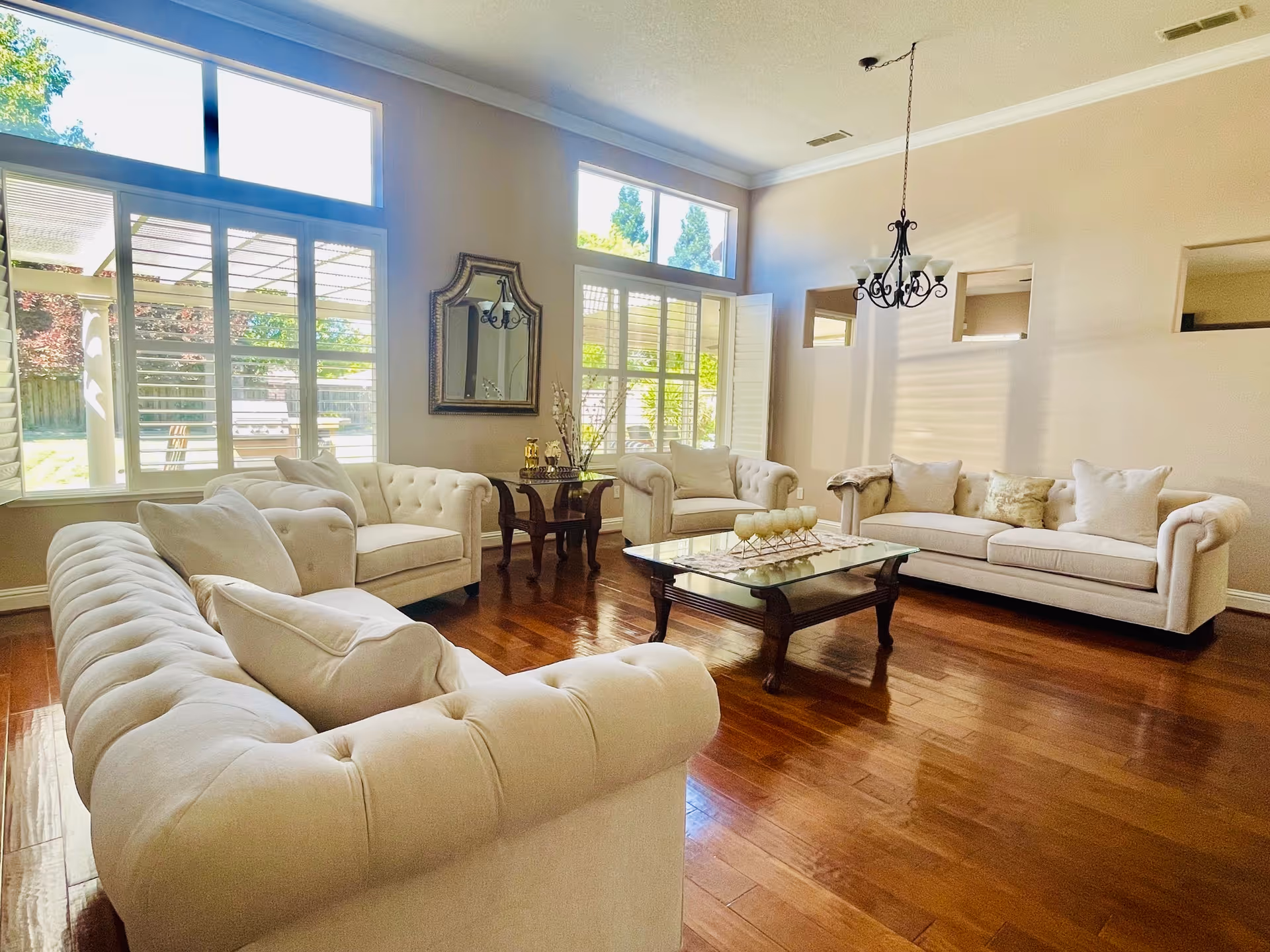 A bright and spacious living room with large windows allowing natural light to fill the space. The room features three cream-colored tufted sofas arranged around a wooden coffee table with a glass top. There is a decorative mirror on the wall and a chandelier hanging from the ceiling. The floor is polished hardwood, and outside the windows, a garden with greenery is visible.