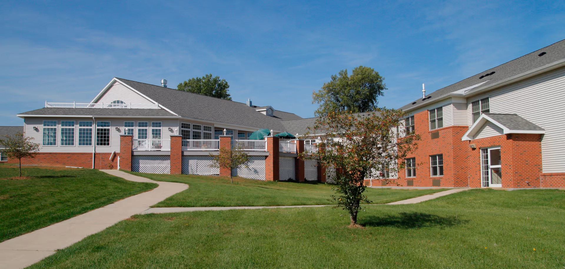 Exterior front view of a brick-and-white senior living facility with a grassy lawn, paved walkways, and a clear blue sky.